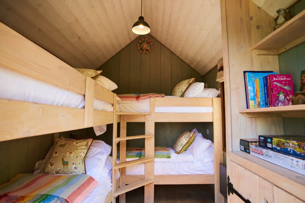 A cozy Dittisham Hideaway cabin room with wooden bunk beds on either side. Each bed is neatly made with colorful quilts and pillows. A warm light hangs from the ceiling, and a clock decorates the wall. Shelves hold books and board games, perfect for a Shepherds Lodges retreat.