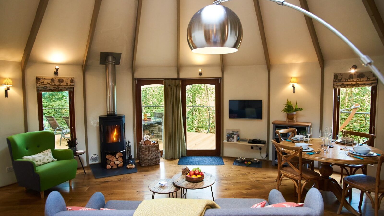 Cozy living room with a high ceiling and circular layout reminiscent of treehouses. Features a fireplace, green and gray sofas, and a dining area with a wooden table and chairs. Large windows open to a deck offering forest views, akin to the serene charm of Dittisham Hideaway. Modern decor, soft lighting.