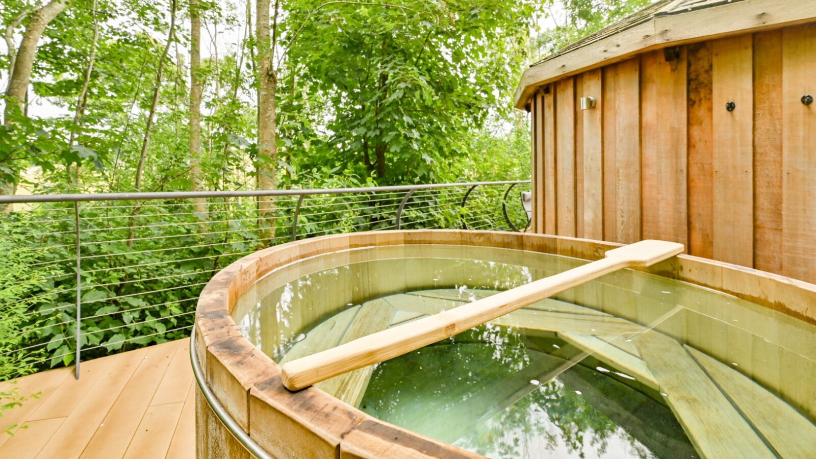 A wooden hot tub filled with water sits invitingly on a deck, enveloped by lush greenery and trees. The tub, adorned with a wooden paddle, is nestled near a charming treehouse at the serene Dittisham Hideaway, promising a perfect escape into nature.