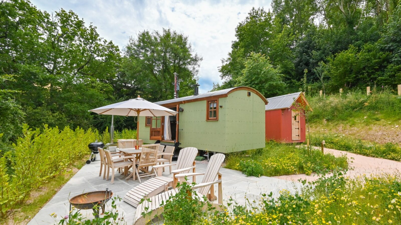 A small, rustic wooden cabin with a curved roof sits surrounded by lush greenery, akin to the cozy Shepherds Lodges at Dittisham Hideaway. In front, patio chairs and a table with a white umbrella grace the stone terrace by the fire pit. The setting is serene and picturesque.