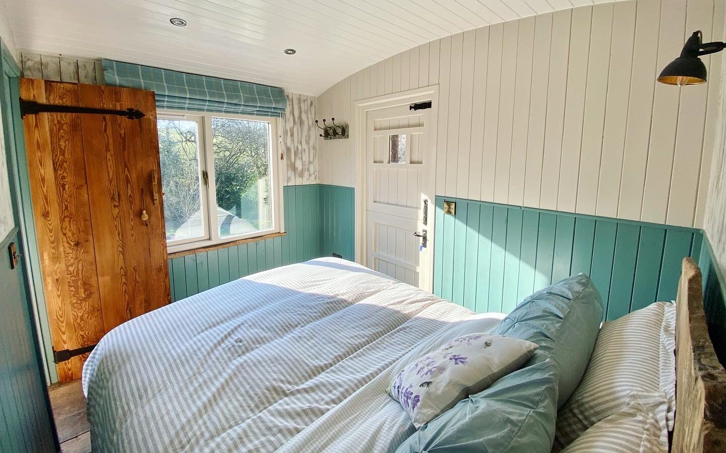 A cozy bedroom featuring a double bed and Caban Eric-inspired blue and white decor, complemented by a wooden door. A window framed by trees invites natural light to dance across the white panel ceiling, creating an inviting retreat.