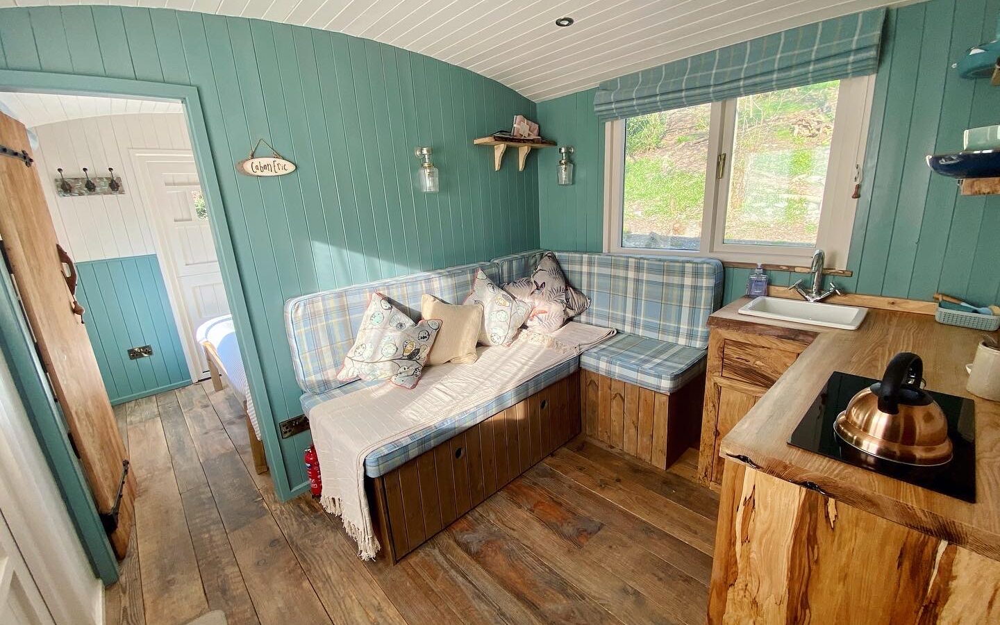 The cozy Caban Eric interior features teal walls, a plaid-cushioned bench, wooden floors, and a quaint kitchenette. A window with a view completes this charming retreat.