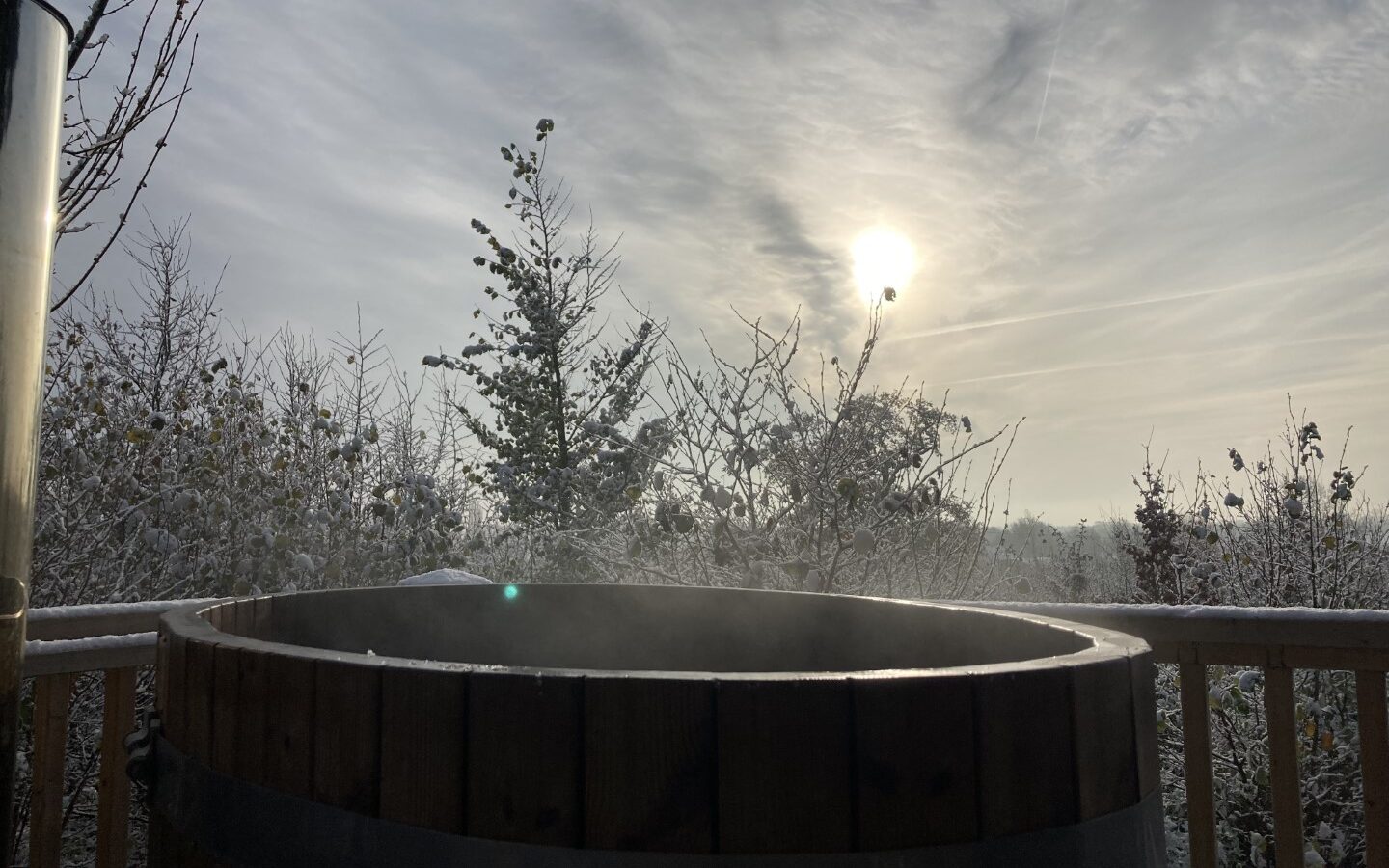 A wooden hot tub on the deck of Abbey Farm overlooks a snowy landscape with bare trees. Steam rises under a cloudy sky, and the sun is partially visible, casting a soft light over the serene scene.