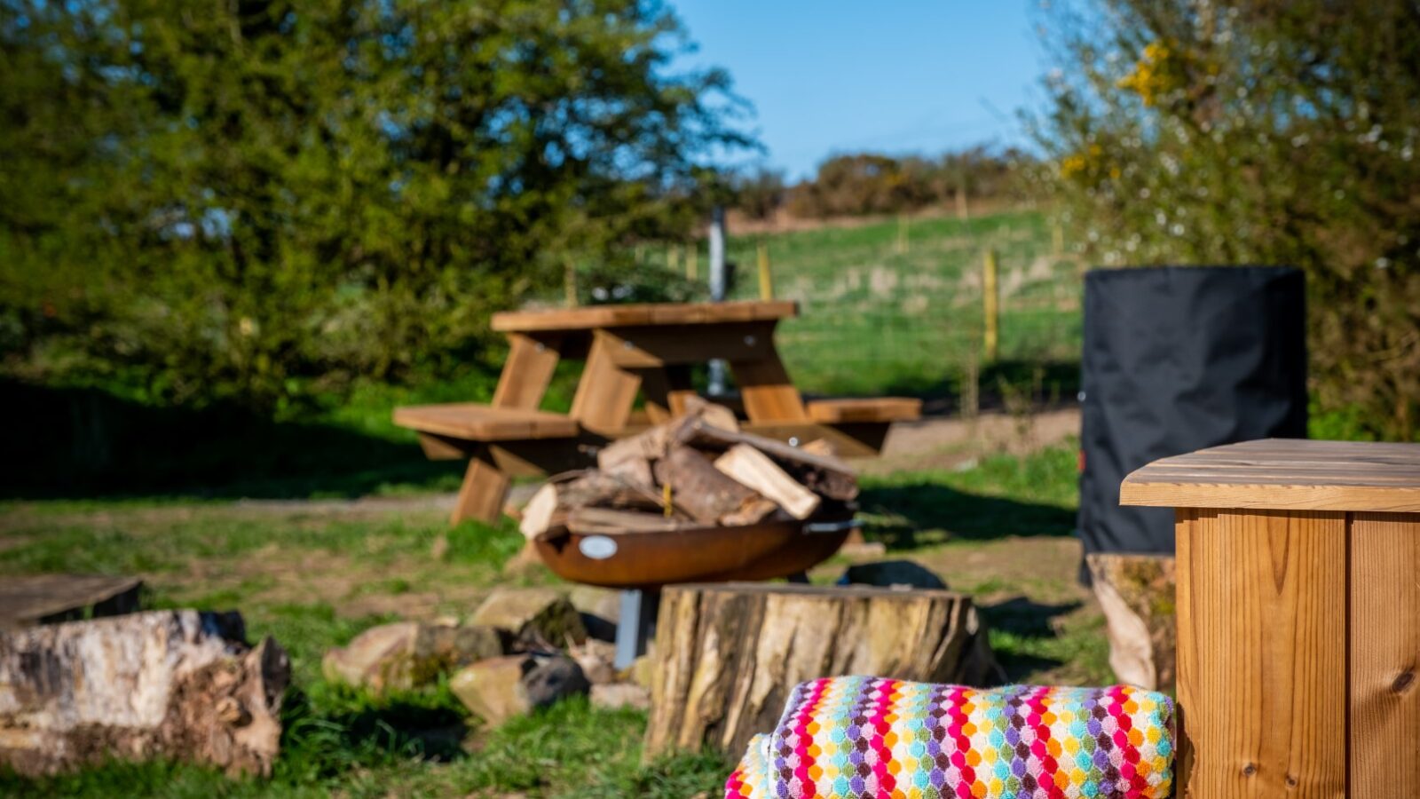 A wooden picnic table with stacked firewood sits in a rustic outdoor setting at Sloeberry Farm, a colorful blanket in the foreground and trees providing a scenic backdrop.