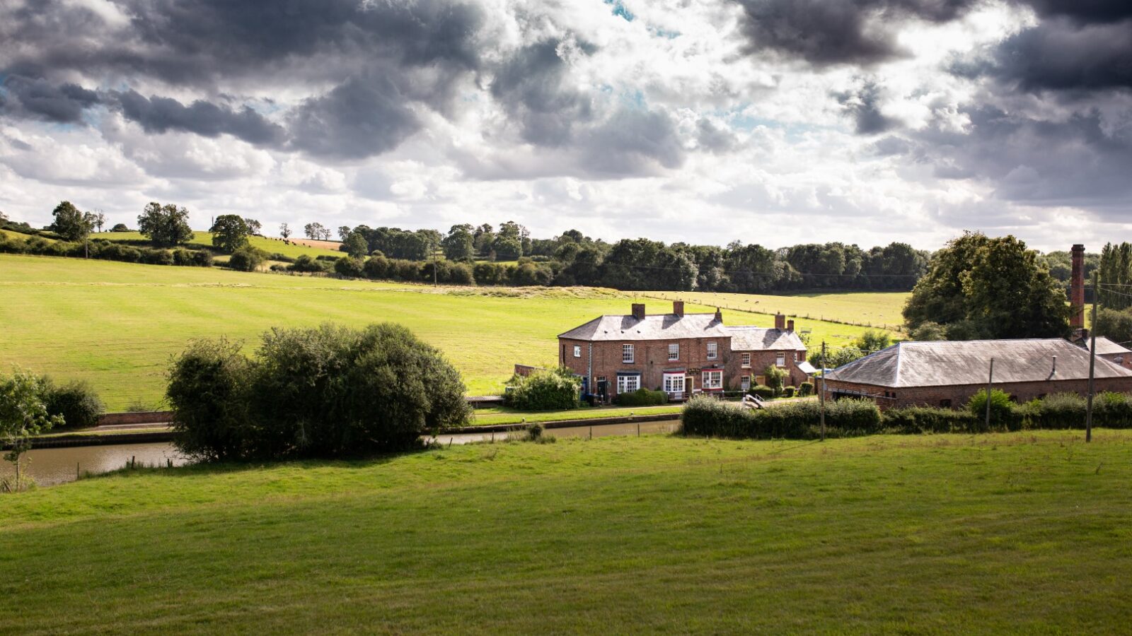 A rural landscape featuring a row of brick houses near a canal, surrounded by lush green fields ideal for Ewe Glamping. The sky is overcast with dramatic clouds, casting shadows on the inviting landscape.