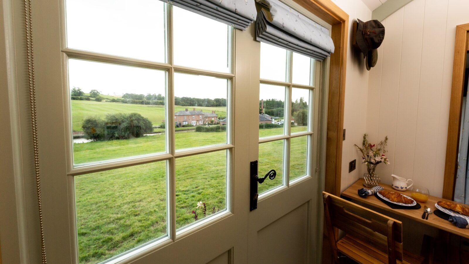 A cozy room with a small wooden table set for breakfast, next to a door with large glass panes. The view outside reveals the picturesque countryside of Ewe Glamping, showcasing green fields, a distant house, and a tree-lined horizon.