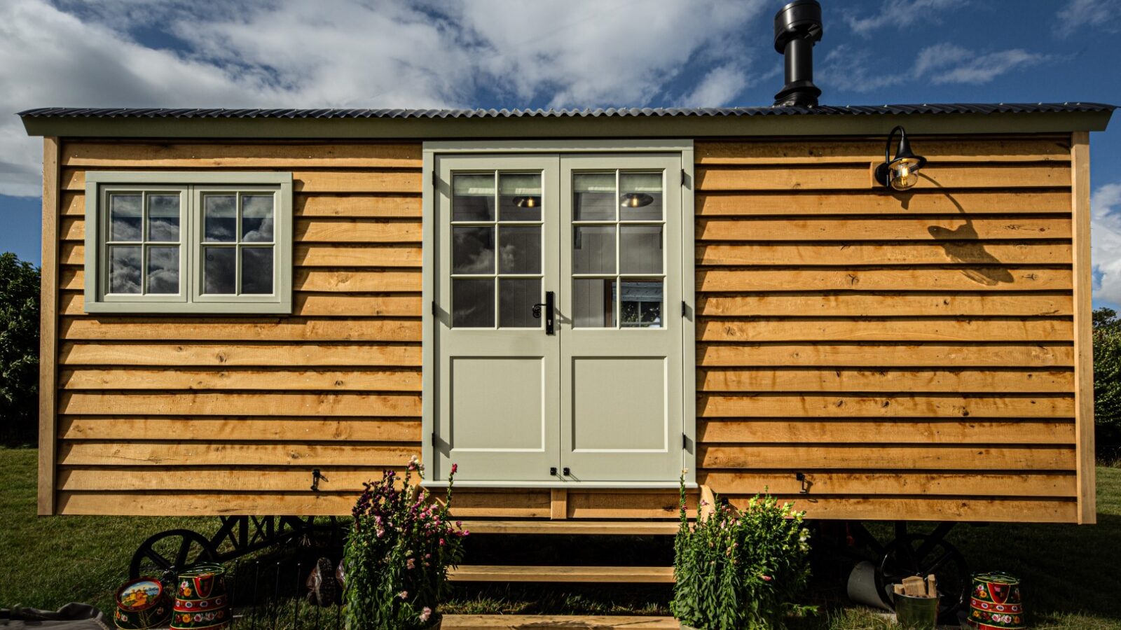 A rustic, wooden tiny house on wheels under a partly cloudy sky offers a delightful glamping experience. With white-framed windows and doors, a charming chimney, and potted plants adorning the front steps, it's an ideal retreat nestled like a peaceful ewe in the countryside.
