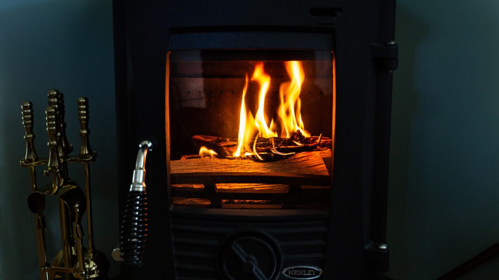 A cozy wood-burning stove with a glass door reveals vibrant orange flames and glowing logs inside, capturing the essence of glamping. A set of brass fireplace tools stands next to it on the left, while the stove emits a warm, inviting glow.