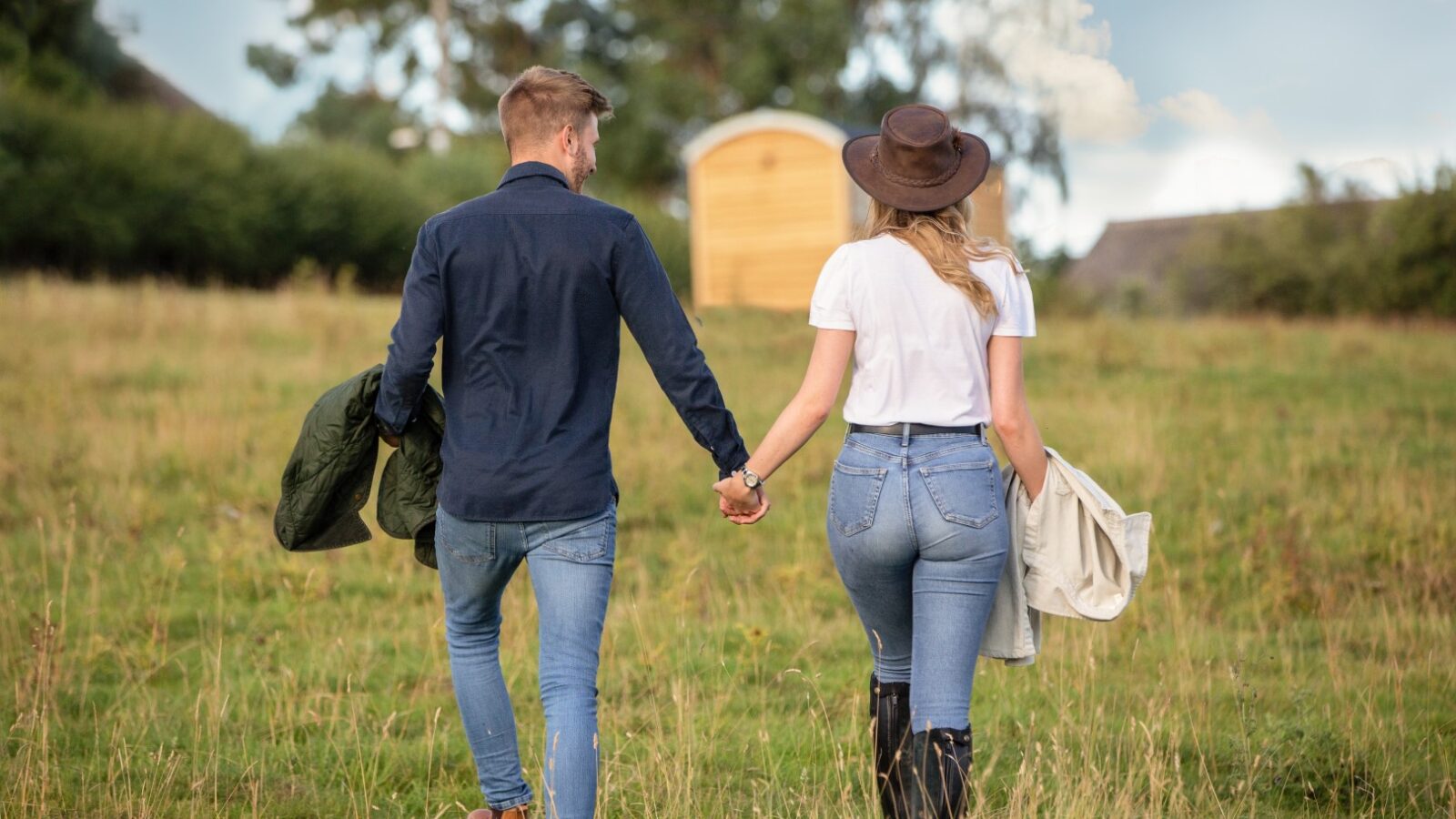 A couple holding hands walks through a grassy field, the man in a dark blue shirt and jeans, carrying a jacket. The woman sports a white shirt, jeans, and a hat. Nearby, a small wooden hut hints at glamping adventures under the stars.