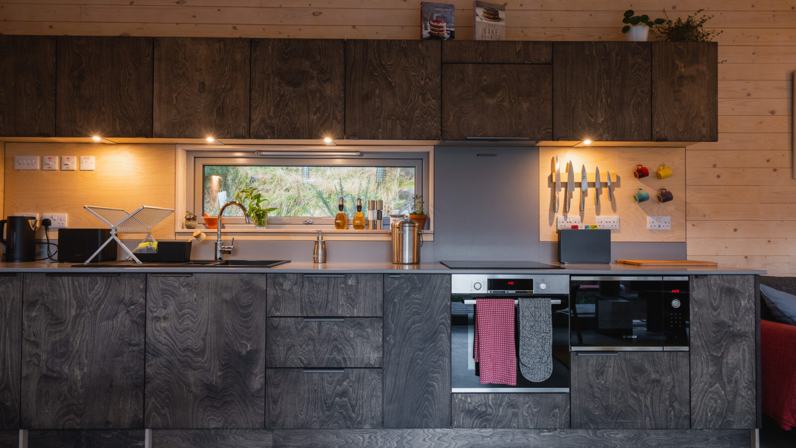 A modern kitchen in an Ecotone Cabins style, featuring wooden cabinets, under-cabinet lighting, a built-in oven, and a stovetop. Knives on a wall magnetic strip complement the dish rack by the sink, with plants atop the cabinets and a window offering an outside view.