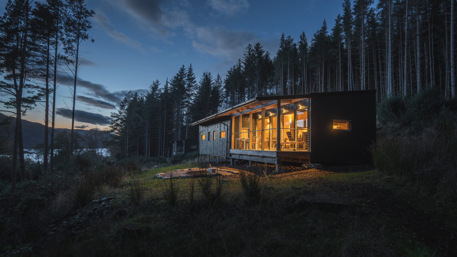 A modern cabin with large windows glows warmly in the evening light, nestled in the ecotone between towering pine trees and a deep blue sky streaked with clouds. In the distance, a shimmering glimpse of water adds to the serene landscape.