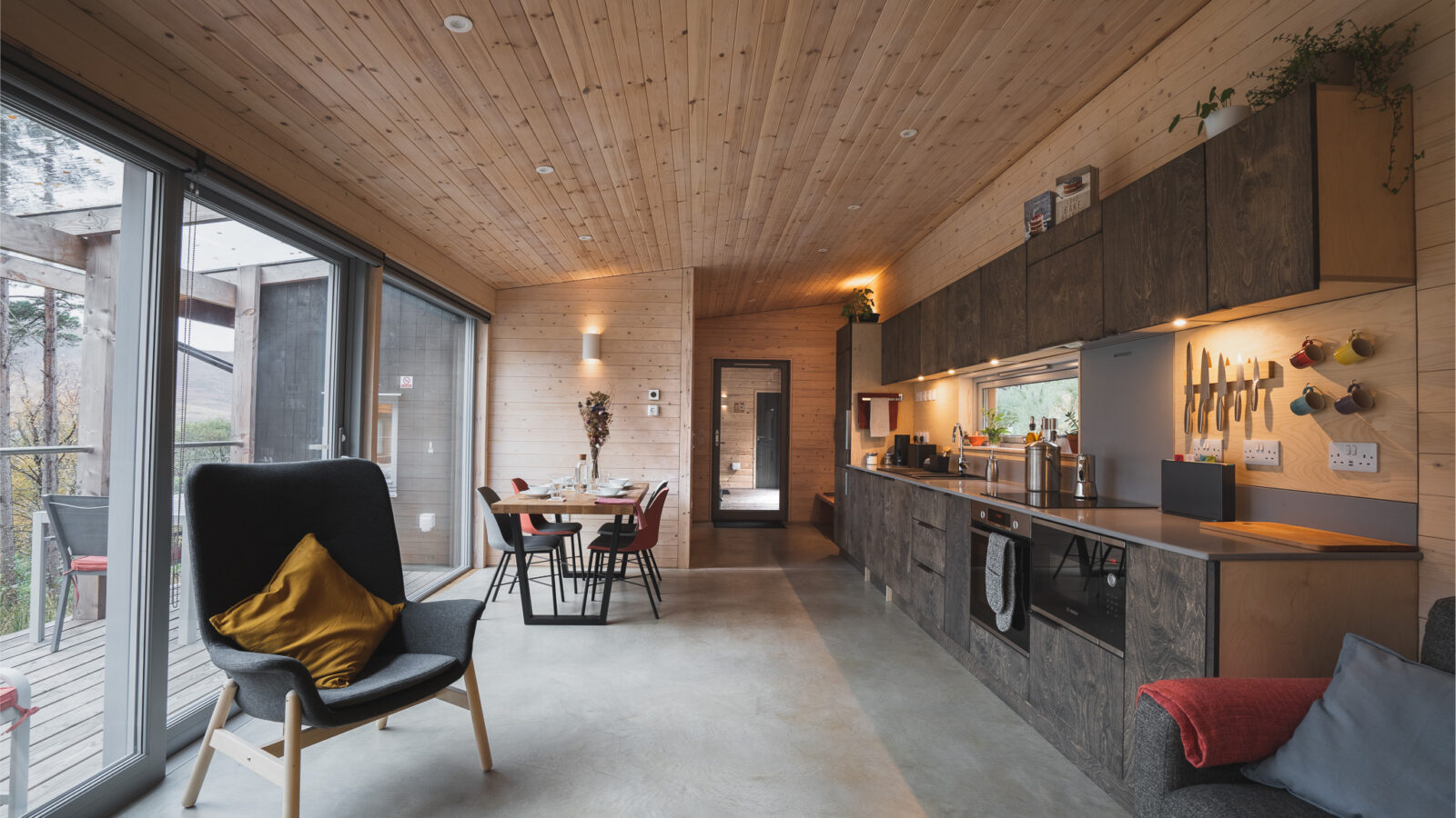 This Ecotone Cabins-inspired modern living space boasts wooden ceilings and walls, featuring a black armchair with a mustard pillow. A dining table complements the kitchen's dark cabinets, while large windows flood the room with natural light and stunning views.