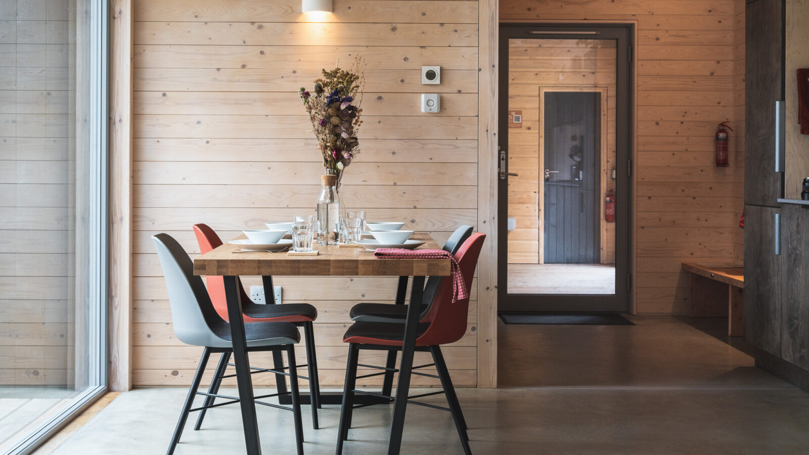 A modern dining room in the style of Ecotone Cabins features a wooden table set for four, with two gray and two red chairs. A vase with dried flowers serves as a centerpiece. The walls and floor are wood-paneled, and a door with glass panes is visible in the background.