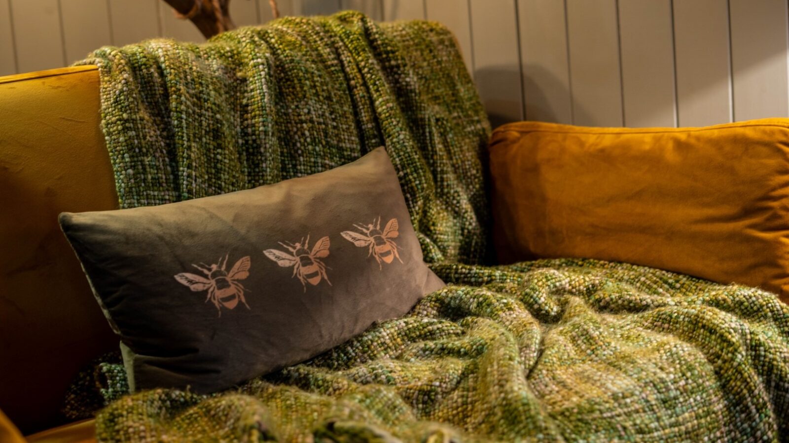 A cozy scene from Erwain Escapes featuring a mustard yellow couch adorned with a green, textured throw blanket. On the couch rests a gray pillow decorated with illustrations of bees. The background consists of light-colored, vertical cabin-style wooden paneling.