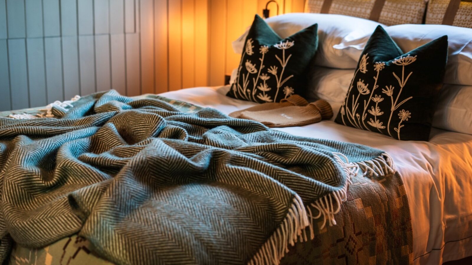 A cozy bedroom at Erwain Escapes features a neatly made bed adorned with green and white patterned pillows and a herringbone throw blanket. Warm lighting creates a comfortable ambiance, and fairy lights in the background add a touch of charm, perfect for your cabin getaway.