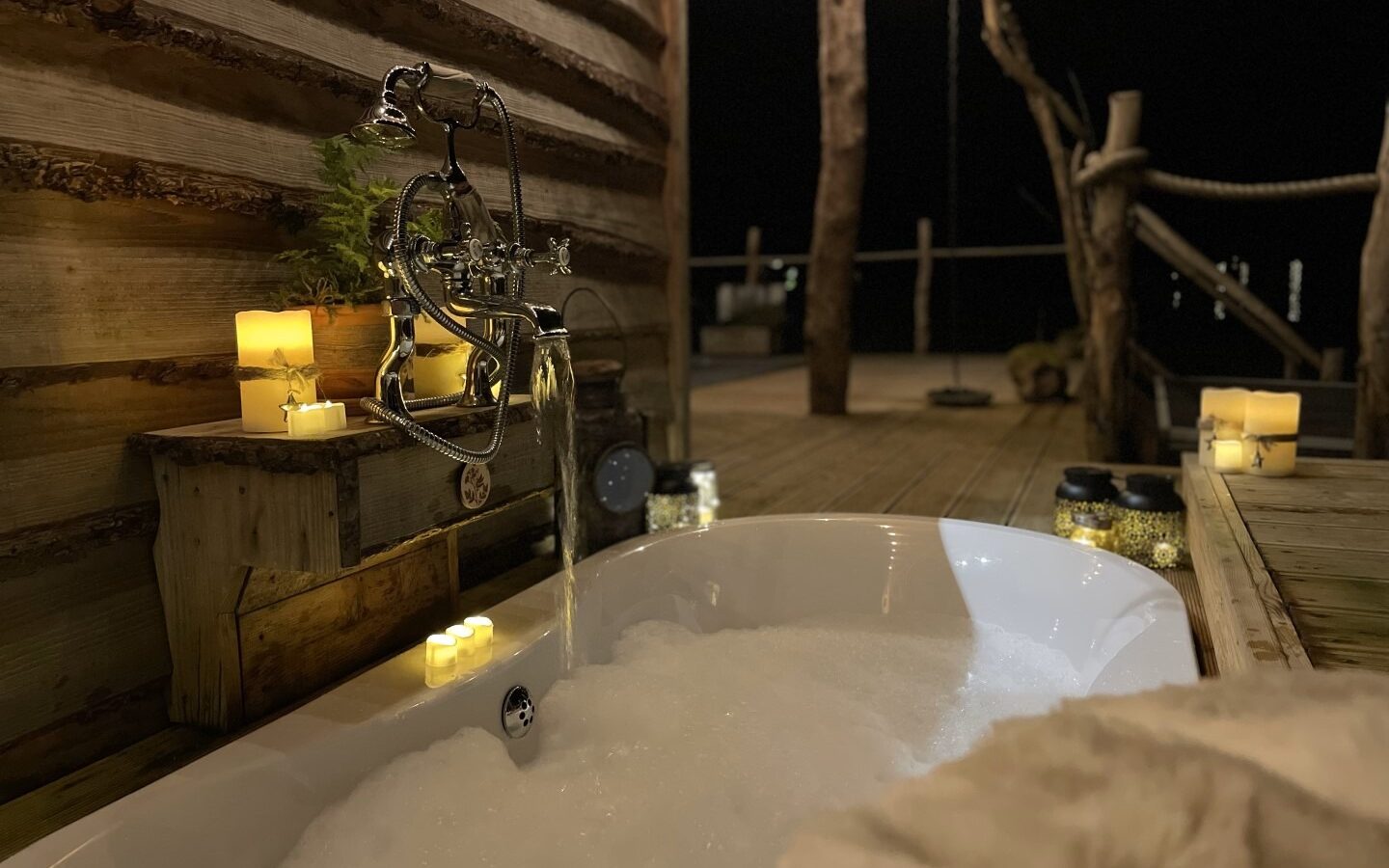 A cozy outdoor bathtub filled with bubbles is set on a wooden deck at night. Illuminated by soft candlelight, a rustic wooden wall adds to the charm. The atmosphere is warm and inviting, perfect for a relaxing soak under the stars at Erwain Escapes' picturesque cabin retreat.