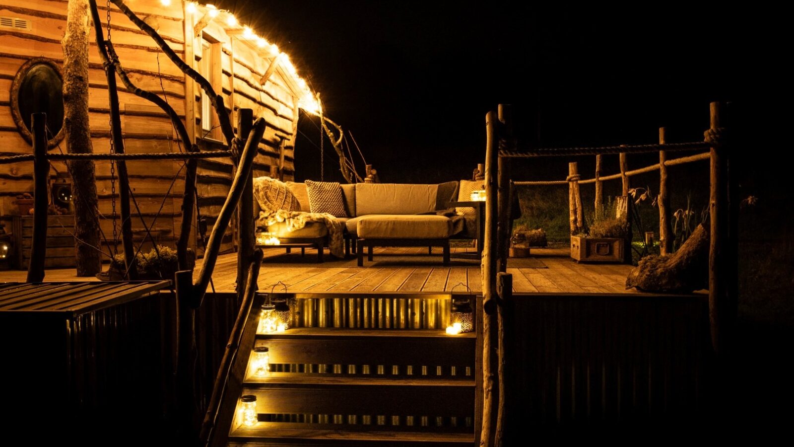 A cozy wooden deck at The Cabin is illuminated by strings of warm fairy lights. A comfortable outdoor seating area with a bench and cushions is set up on the deck. The surrounding darkness enhances the inviting glow of the lights, creating a tranquil nighttime setting at Erwain Escapes.