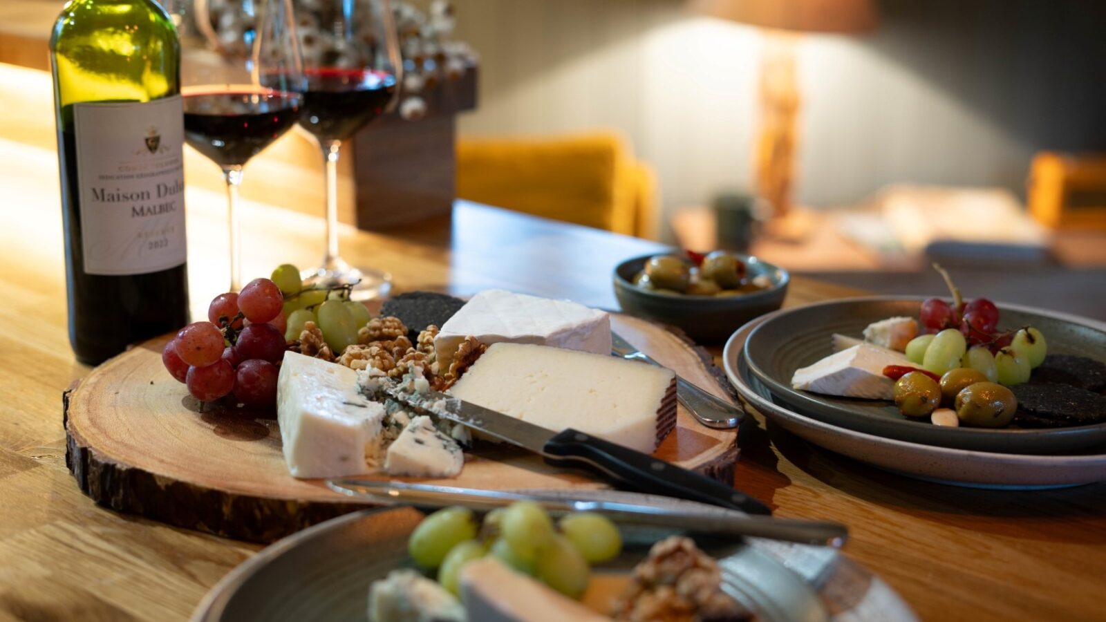 A wooden table with a rustic cheese platter featuring assorted cheeses, grapes, and walnuts at The Cabin. Two glasses of red wine and a bottle labeled 