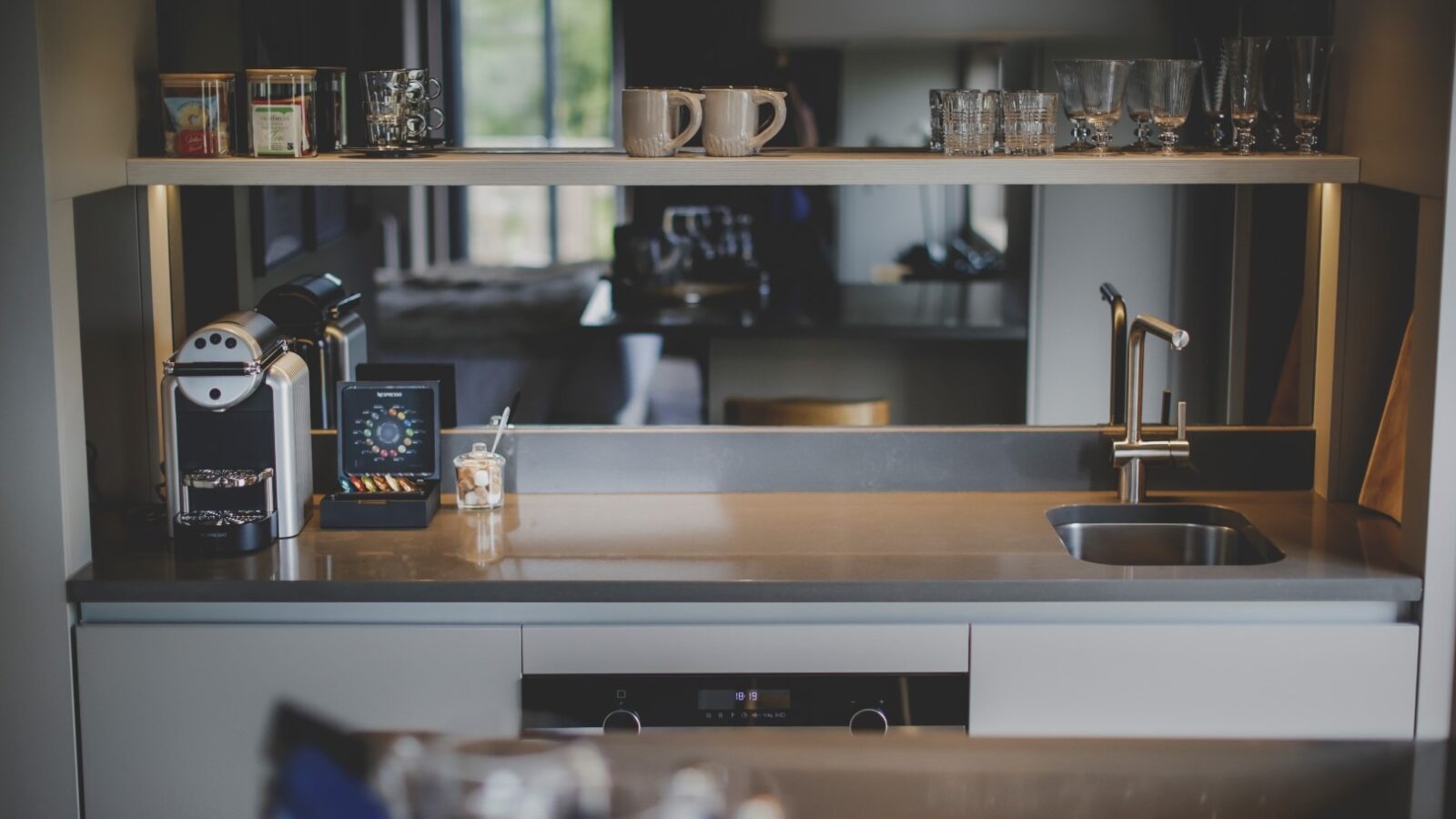 A modern kitchen with a sleek countertop, a sink with a faucet, a coffee machine with pods, and a small jar of utensils. Various glasses and cups are placed on a shelf, creating an inviting retreat. The well-lit space features a window in the background, offering a serene glimpse of greenery outside.