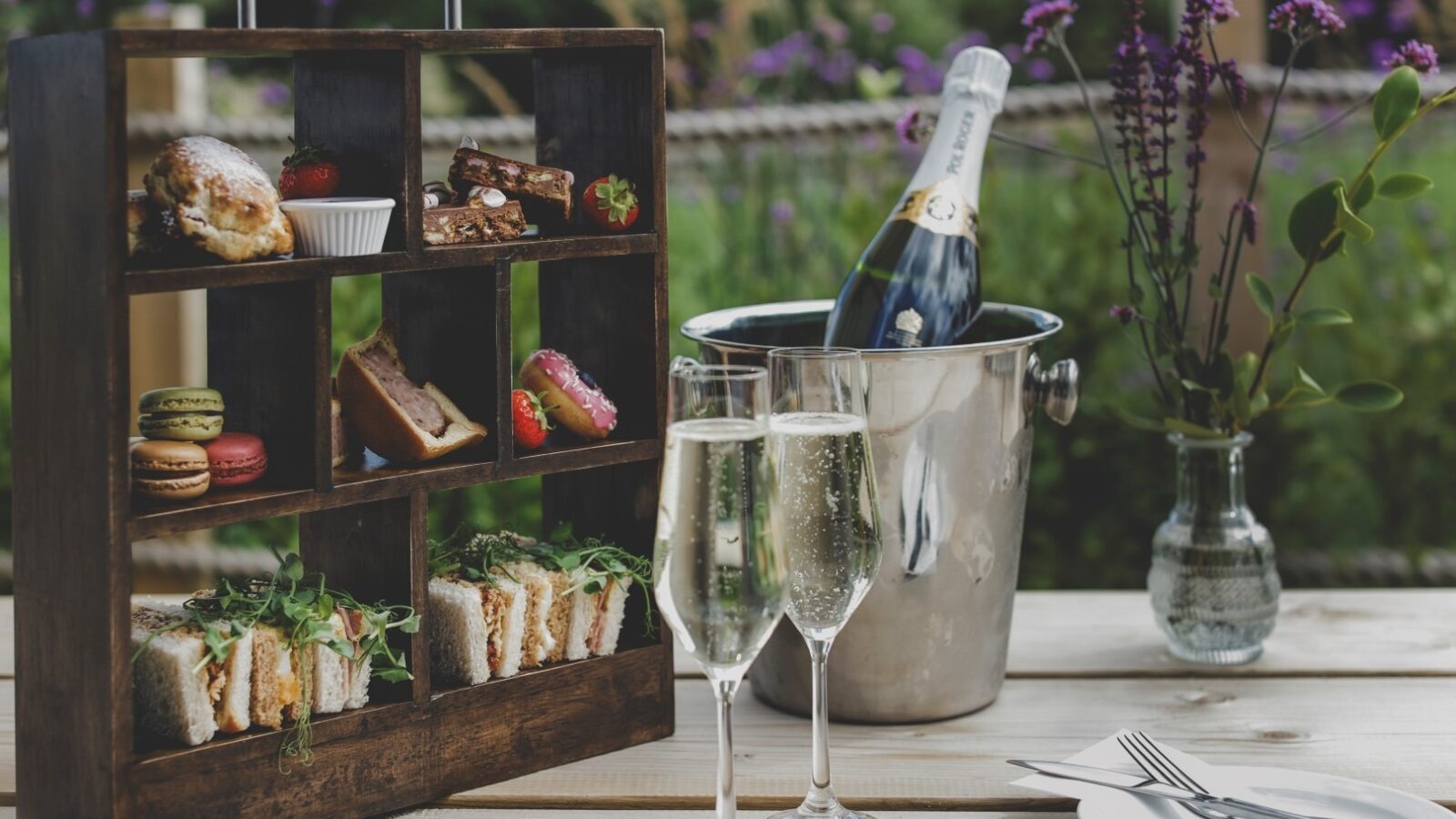 A stylish wooden rack holds an assortment of pastries, macarons, sandwiches, and desserts on an outdoor table—a perfect retreat. Nearby, a bottle of champagne sits in an ice bucket with two filled champagne flutes beside it. A vase with purple flowers completes the elegant setting.