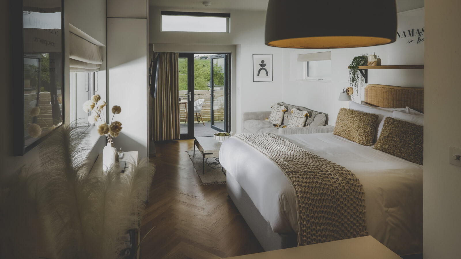 A bright, modern bedroom serves as a perfect retreat, with a large bed adorned with white linens and brown textured pillows. The room features a balcony with a scenic outdoor view, houseplants, a small seating area, and minimalist decorations, creating a serene and cozy atmosphere.