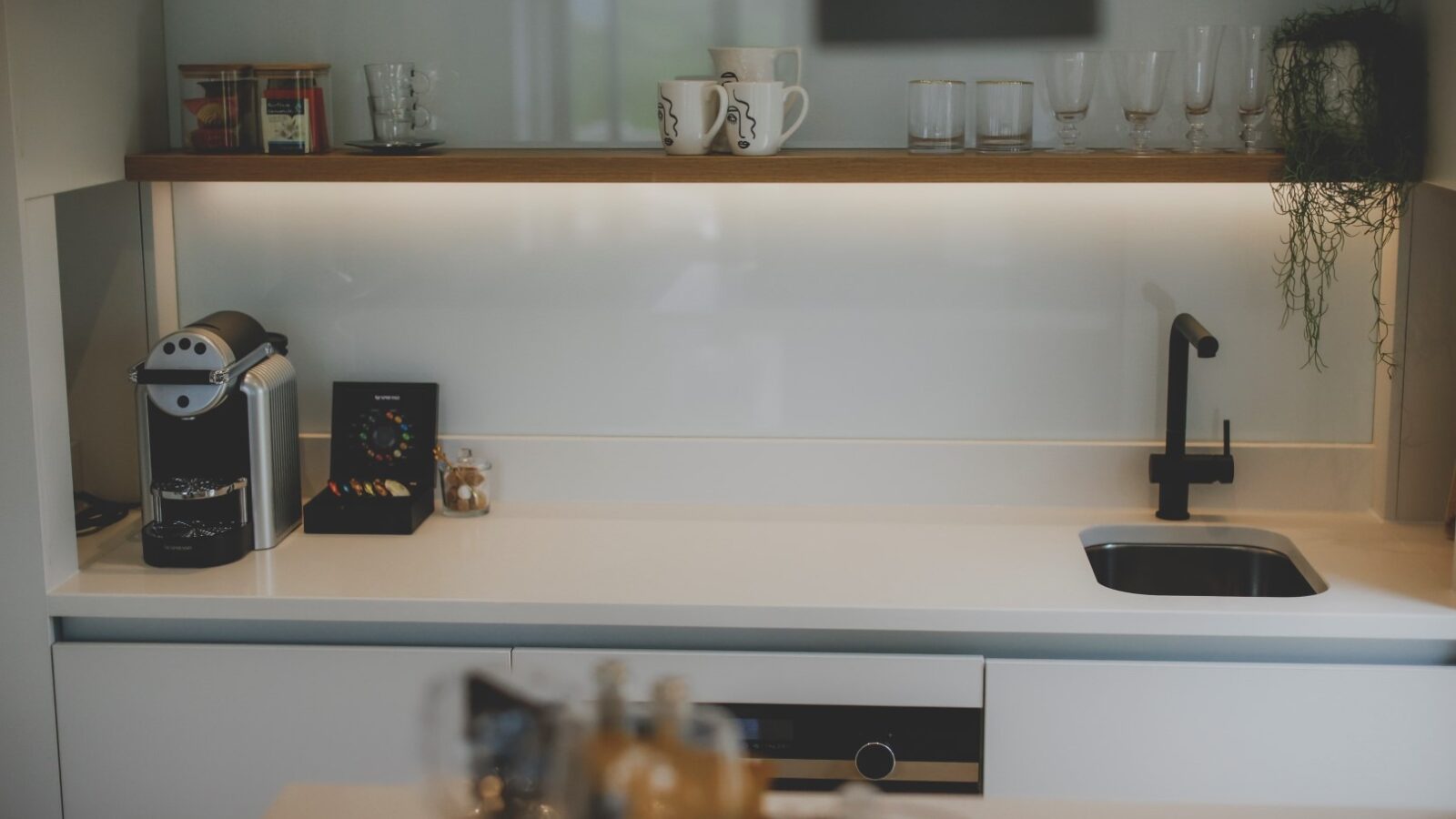 A modern kitchen features a small sink with a black faucet, a coffee machine, and a spice rack on the counter. Shelves above hold glasses, mugs, and plants. A few wine glasses are in the foreground on the counter. The minimalist design of this culinary retreat is highlighted by sleek white surfaces.