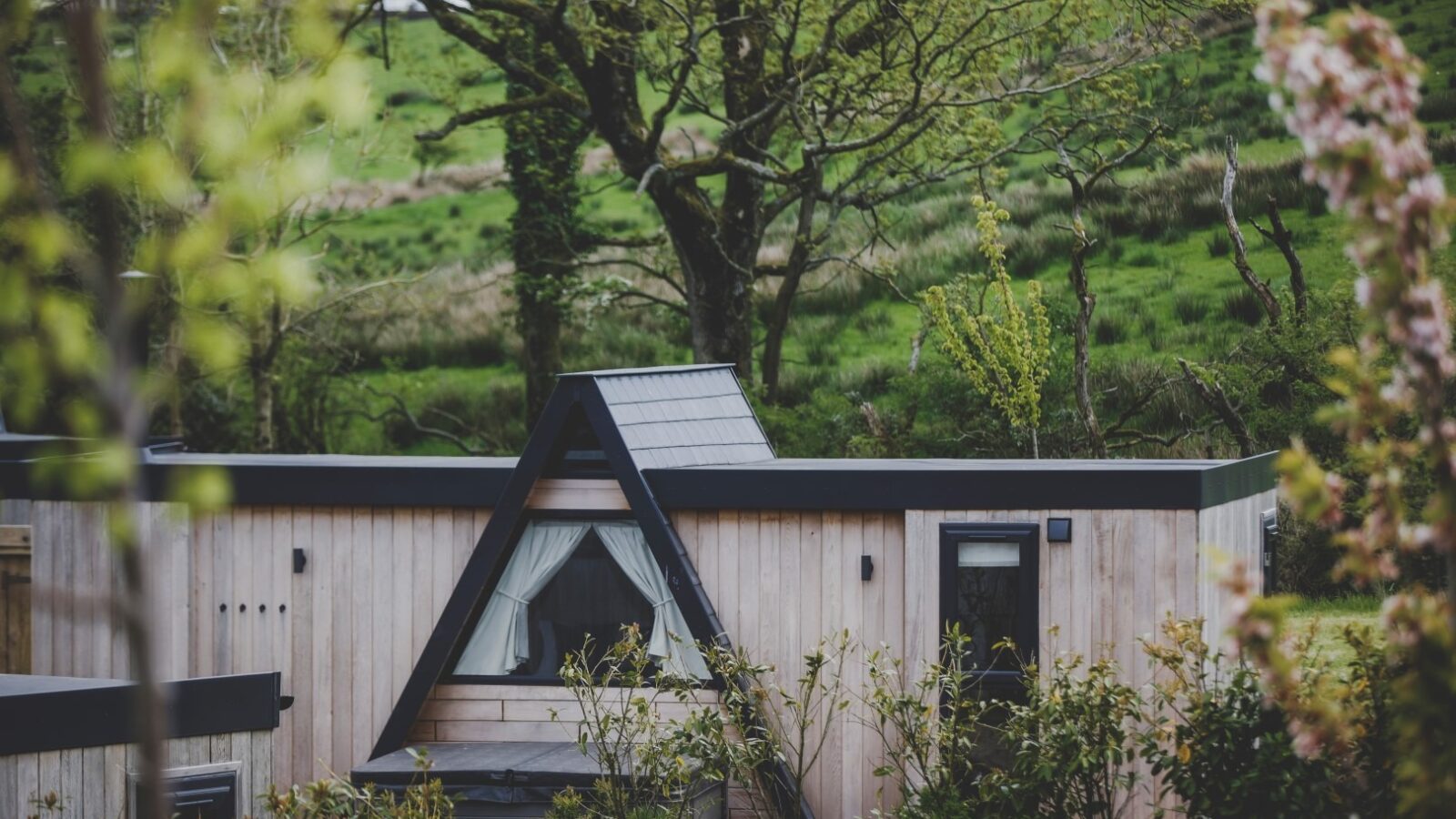 A small, wood-paneled house with an A-frame roof in a lush, green, rural area offers the perfect retreat. The house has white curtains in the triangular window, surrounded by bushes and trees with budding leaves. A scenic background of rolling green hills and more trees is visible.