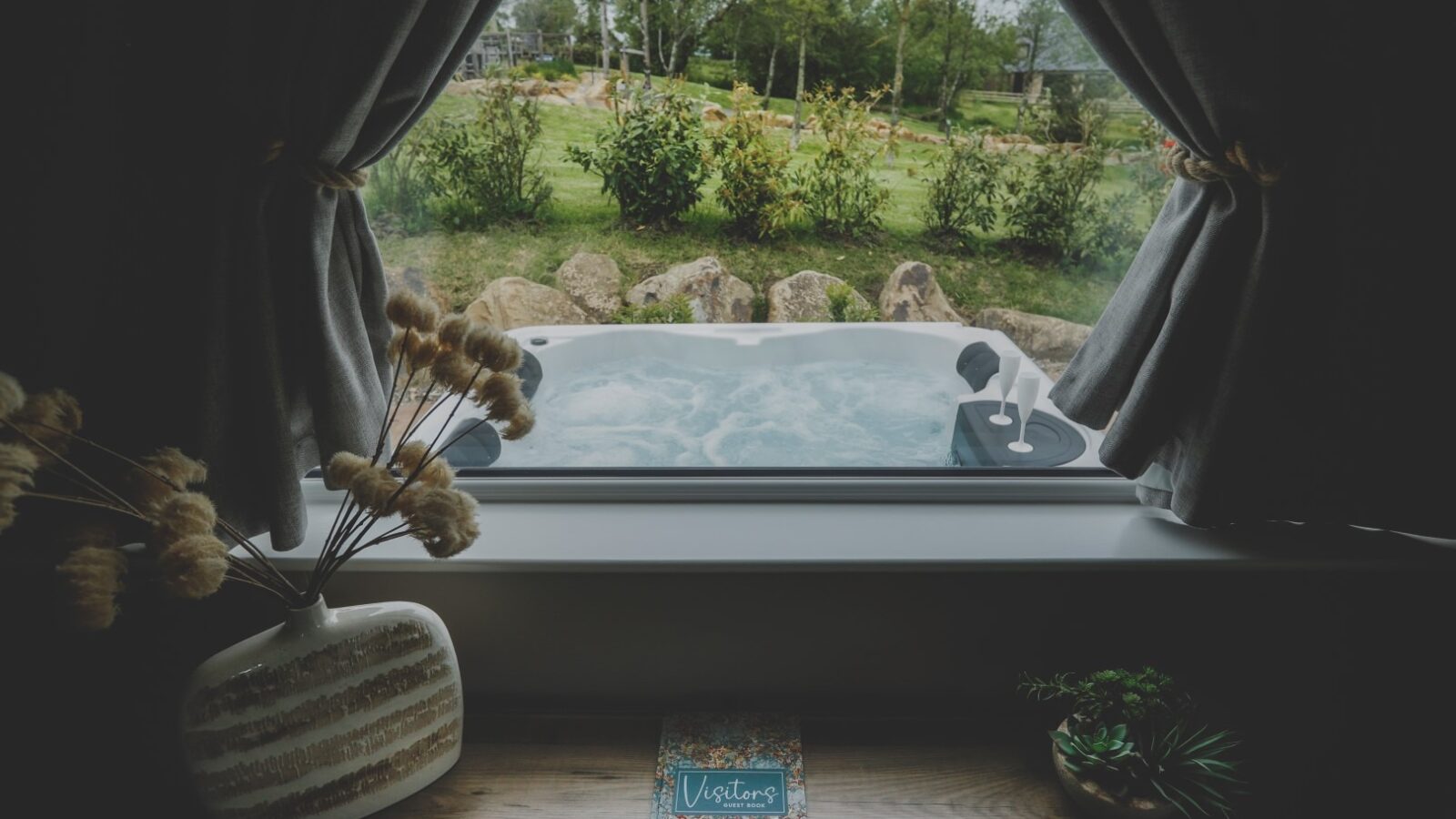 A cozy retreat room with a window view showcasing a bubbling hot tub outside. Curtains frame the view, and a table with a vase of dried flowers and a visitors book is in the foreground. Lush greenery and a garden are visible in the background.