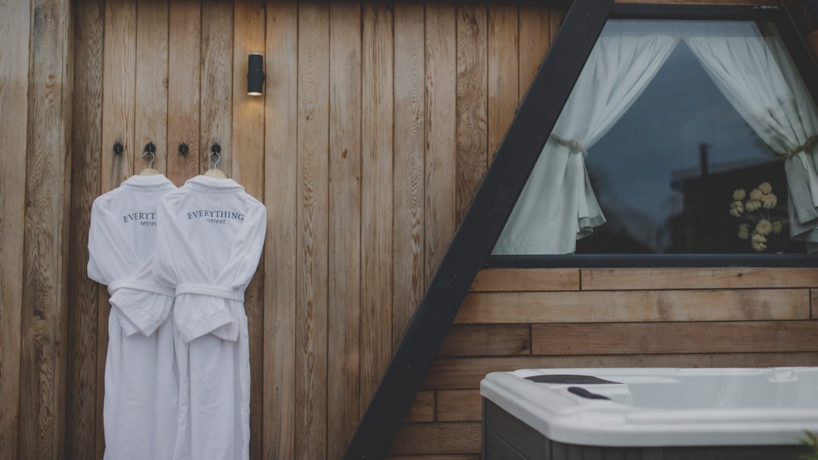 Two white bathrobes hang on hooks attached to a wooden wall of a cabin with a triangular roof, evoking a serene retreat. A light fixture shines above the bathrobes. To the right, a window with white curtains is visible, and at the bottom, part of a hot tub can be seen.