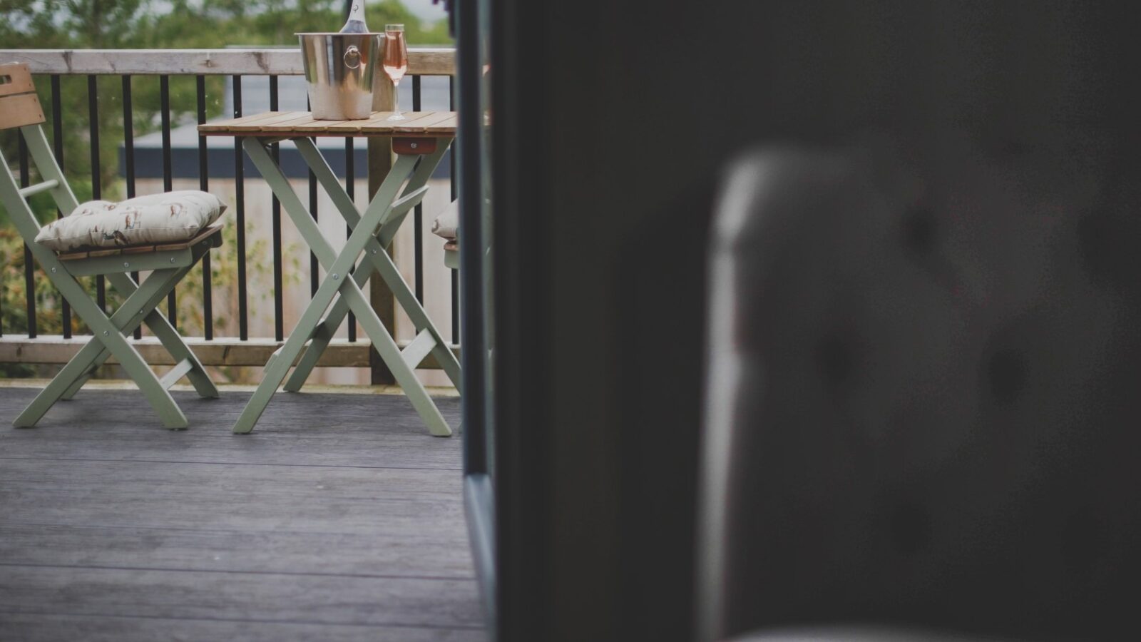 A cozy outdoor patio set, perfect for a relaxing retreat, features two wooden chairs and a small table placed on a wooden deck. On the table, an ice bucket holds a bottle with two glasses filled with rosé wine. The background includes a wooden railing and blurred greenery.