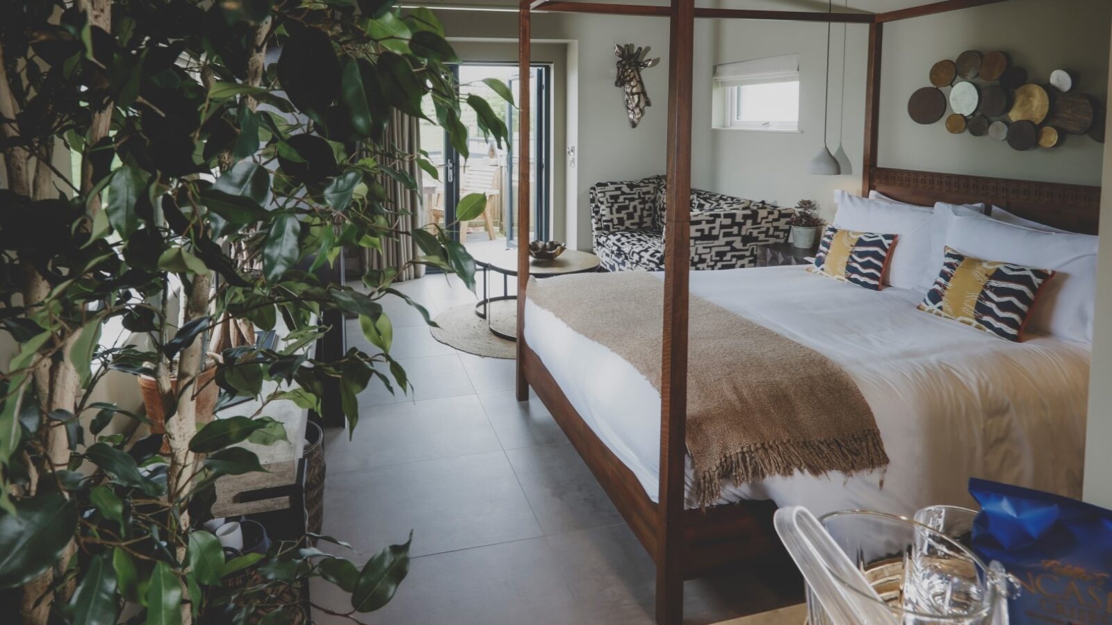 A modern bedroom retreat with a large canopy bed adorned with striped pillows and a beige throw blanket. The room features a potted plant in the foreground, decorative plates on the wall, and a seating nook by a large window offering ample natural light—everything designed for tranquility.