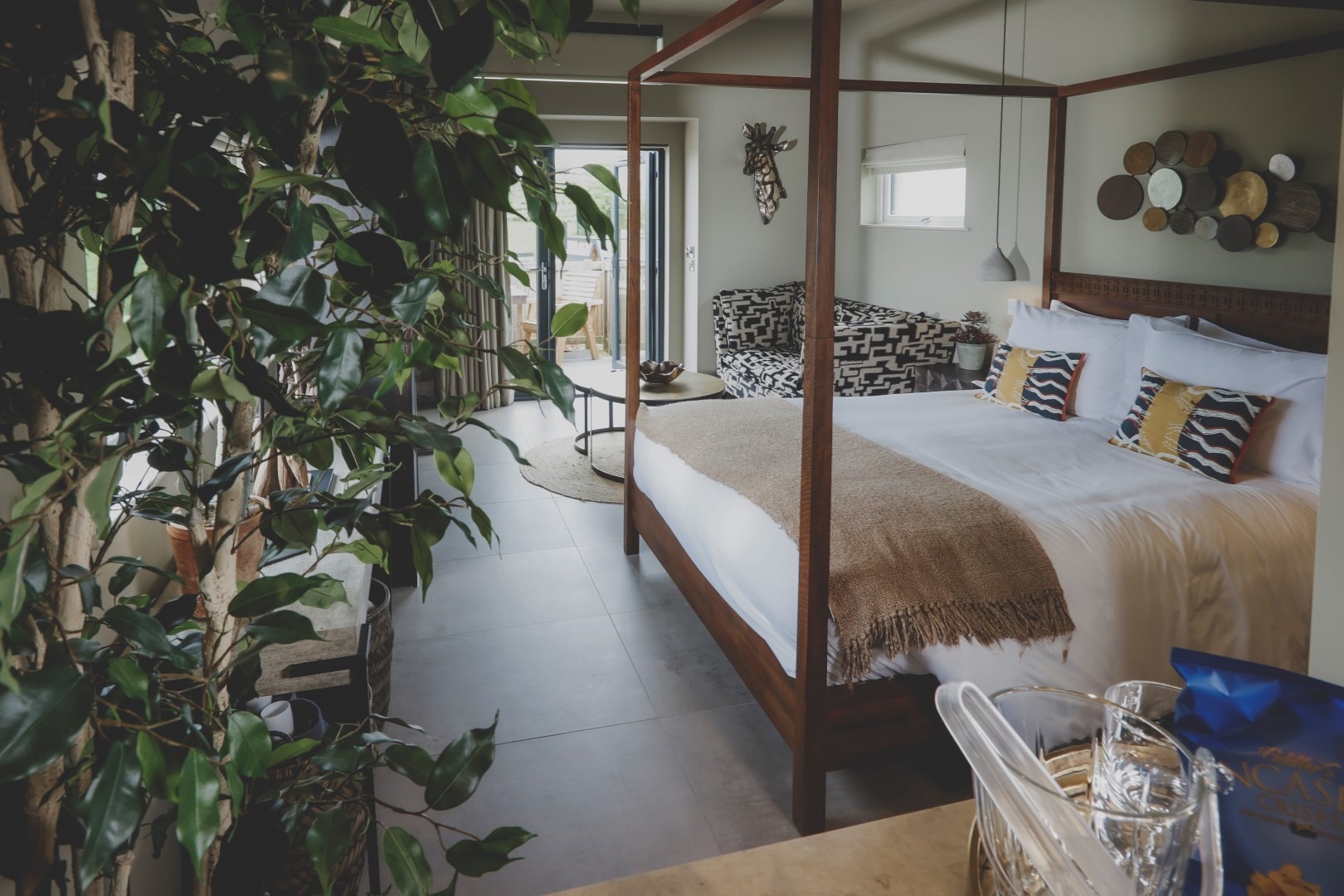 A modern bedroom retreat with a large canopy bed adorned with striped pillows and a beige throw blanket. The room features a potted plant in the foreground, decorative plates on the wall, and a seating nook by a large window offering ample natural light—everything designed for tranquility.