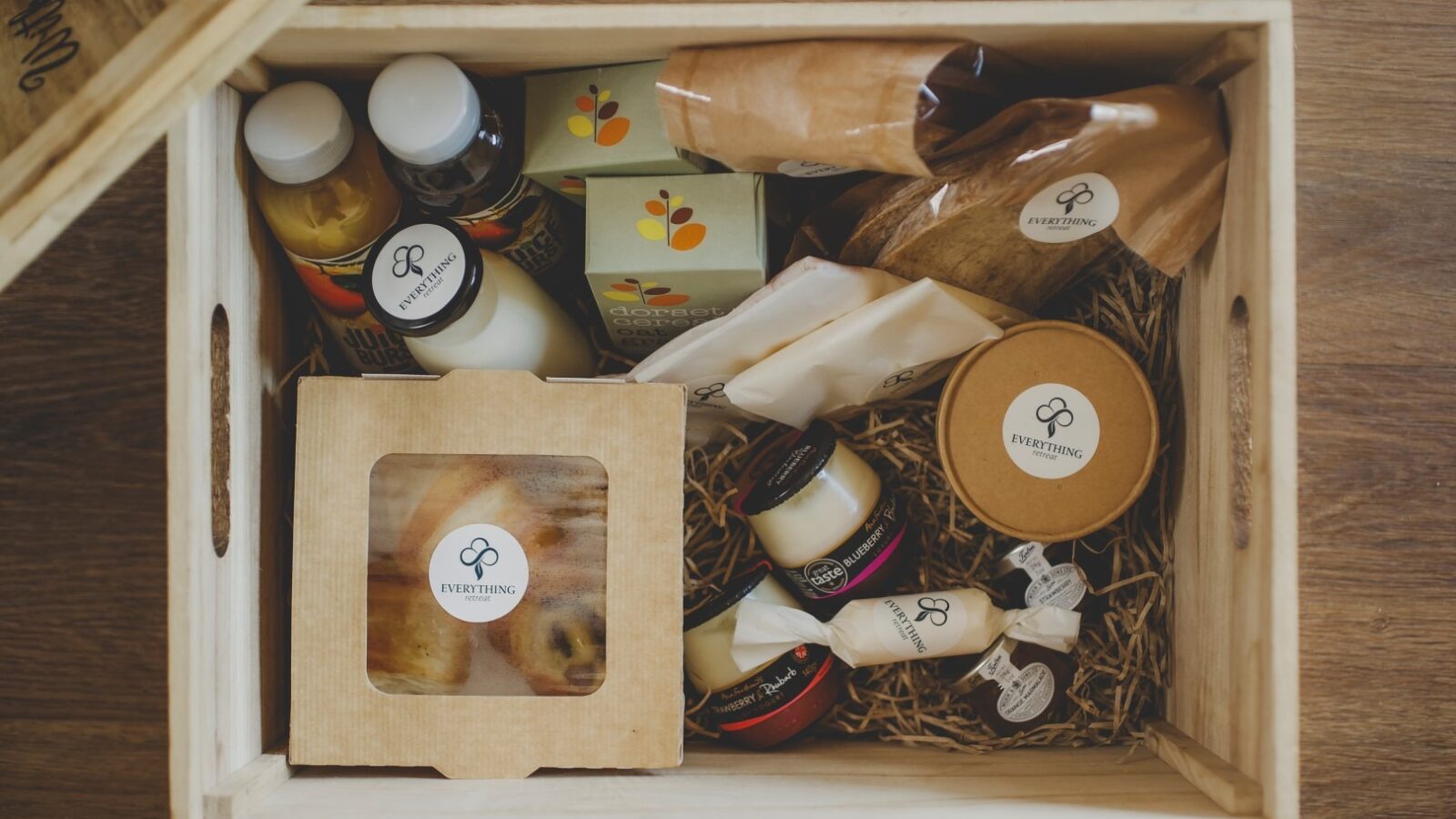 A wooden gift box from Everything Retreat contains various gourmet items, including bottles of dark liquid, jars of honey, cheese, a box with pastries, and packages of tea, nuts, and other treats. The items are neatly arranged with straw-like packaging material.