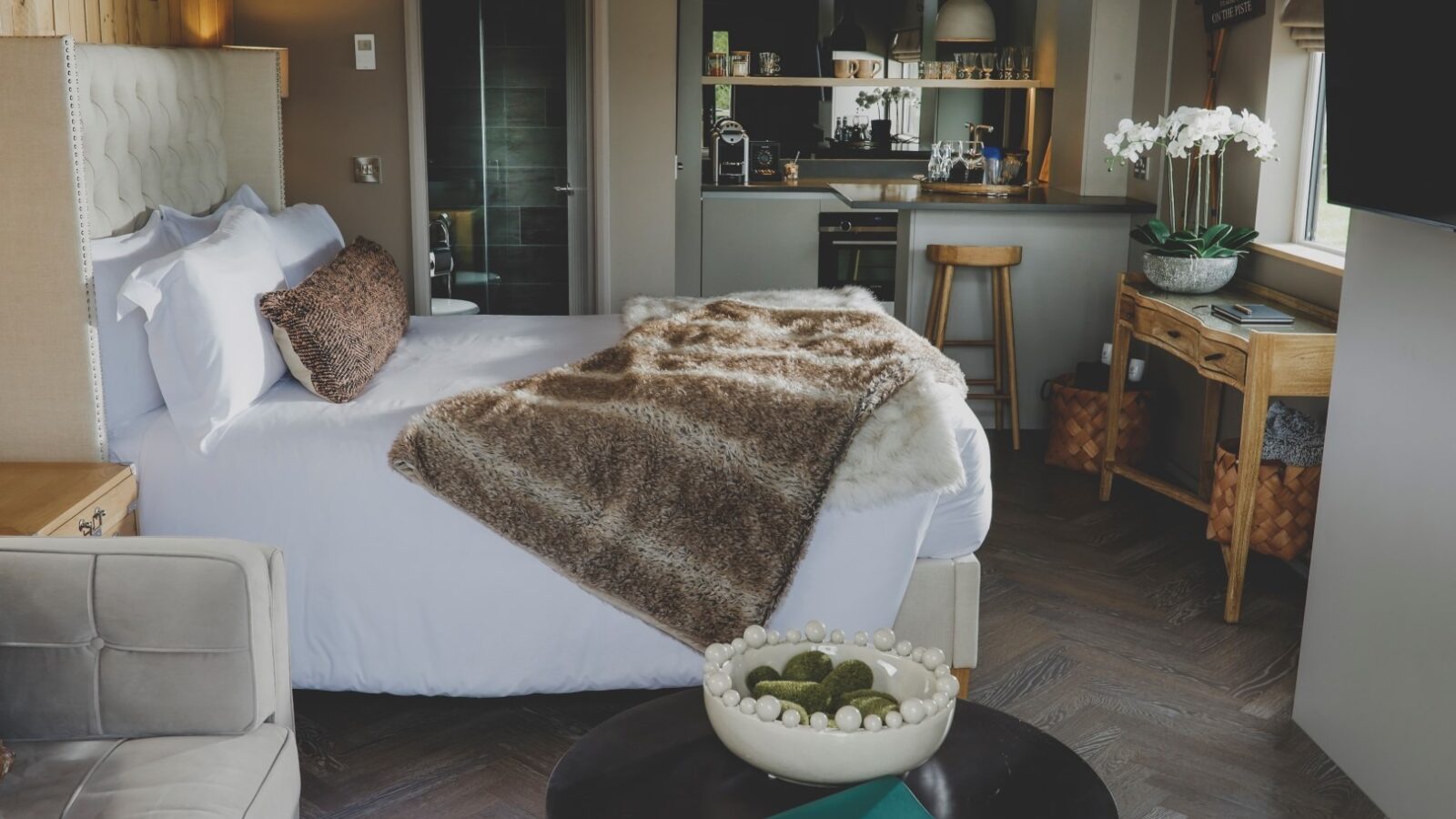 A cozy hotel room, ideal for a retreat, features a plush bed with white linens and a brown faux fur throw. The space includes a small kitchenette, a seating area with decorative items, and a wooden desk adorned with orchids. Warm wooden accents create an inviting atmosphere perfect for everything retreat-related.