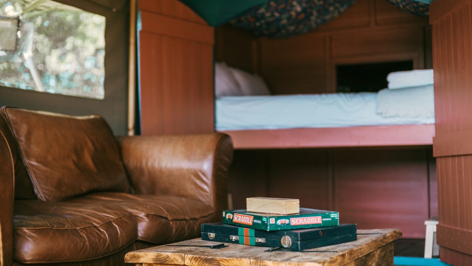 A cozy room in the heart of Exe Valley features a brown leather loveseat, a wooden coffee table topped with Scrabble games and books, and a neatly made bed in the background. The reddish-brown wooden walls and window letting in natural light make it perfect for glamping enthusiasts.