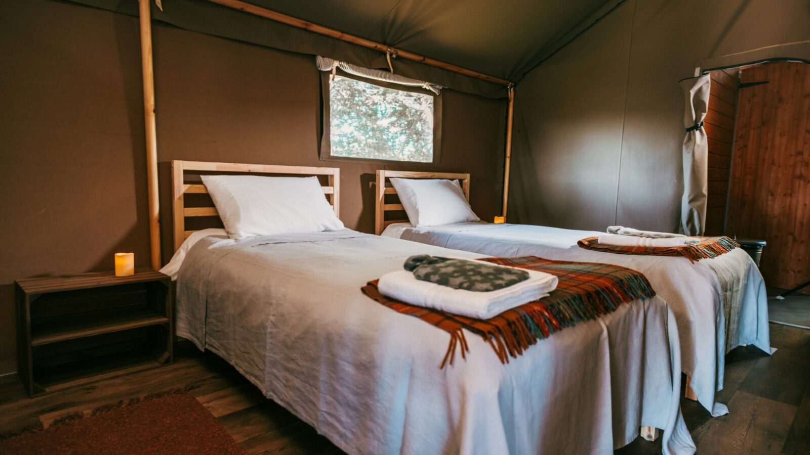 A cozy tent interior at Exe Valley Glamping features two neatly made single beds, each adorned with white linens, folded blankets, and a small candle on a nightstand. The rustic decor includes a wooden floor, a small window, and soft ambient lighting.