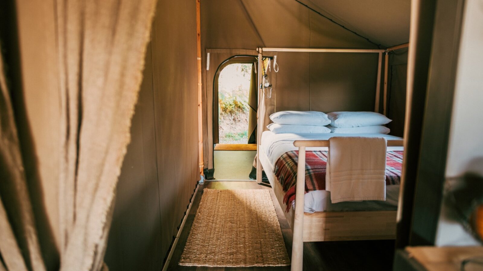 Cozy interior of an Exe Valley Glamping tent with a neatly made bed that has white pillows and blankets, a colorful striped throw blanket, and a folded towel. The open door reveals a glimpse of greenery outside. The tent has wooden flooring and a beige curtain partially drawn to the side.