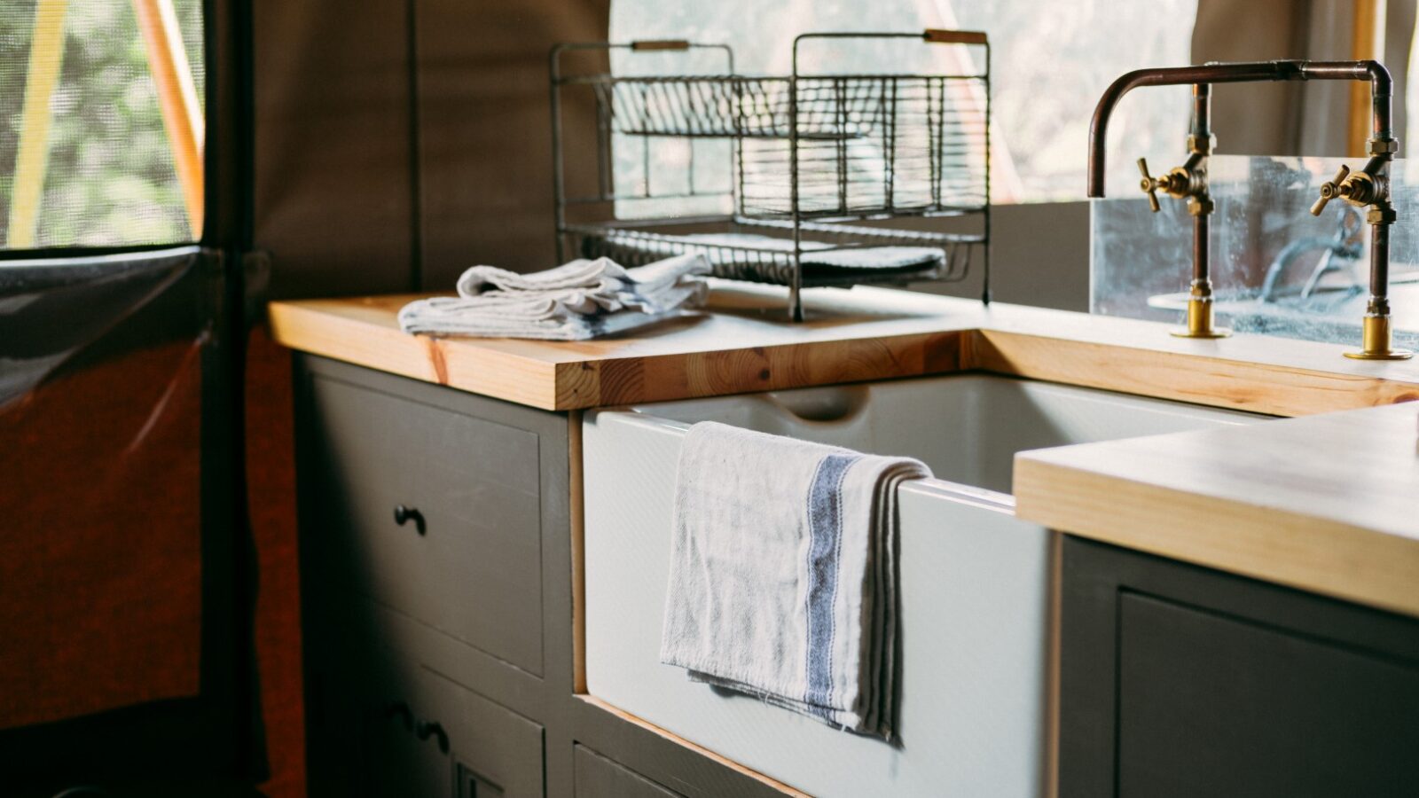 A close-up of a rustic kitchen countertop at Exe Valley Glamping features a farmhouse sink with a towel draped over it. On the counter, there are folded towels and a dish drying rack. Brass faucets are mounted above the sink, and surrounding cabinets boast dark finishes.