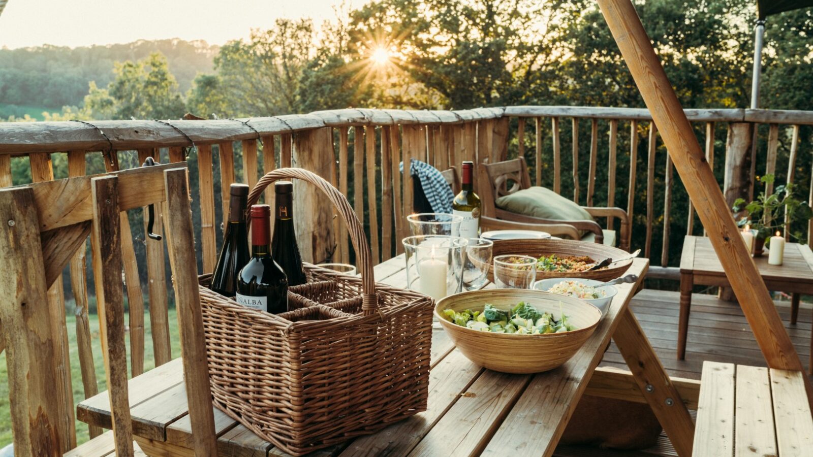 A wooden outdoor picnic table set for a meal. The table holds a wicker basket with wine bottles, glasses, a bowl of salad, bread, and other dishes. The setting is on a wooden deck with a wooden railing, overlooking the scenic Exe Valley with trees and the sun setting—a perfect glamping experience.