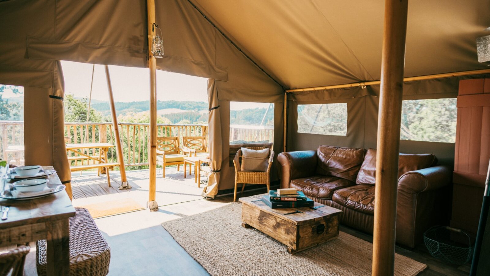 A cozy glamping tent in Exe Valley with a wooden floor, featuring a brown leather sofa, wicker chairs, a rustic coffee table, and a small dining area. The tent opens to a deck with more seating, offering a scenic view of the lush green landscape.