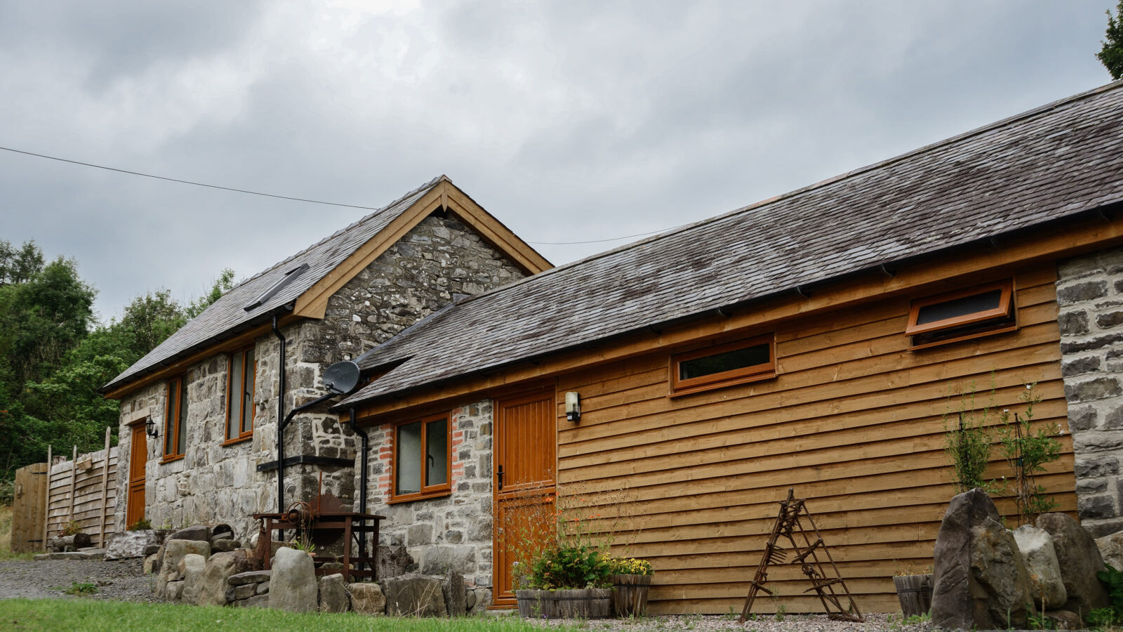 Nant Awen’s charming cottage, crafted from stone and wood, boasts a sloped roof and is nestled amidst lush grass and towering trees under a cloudy sky.