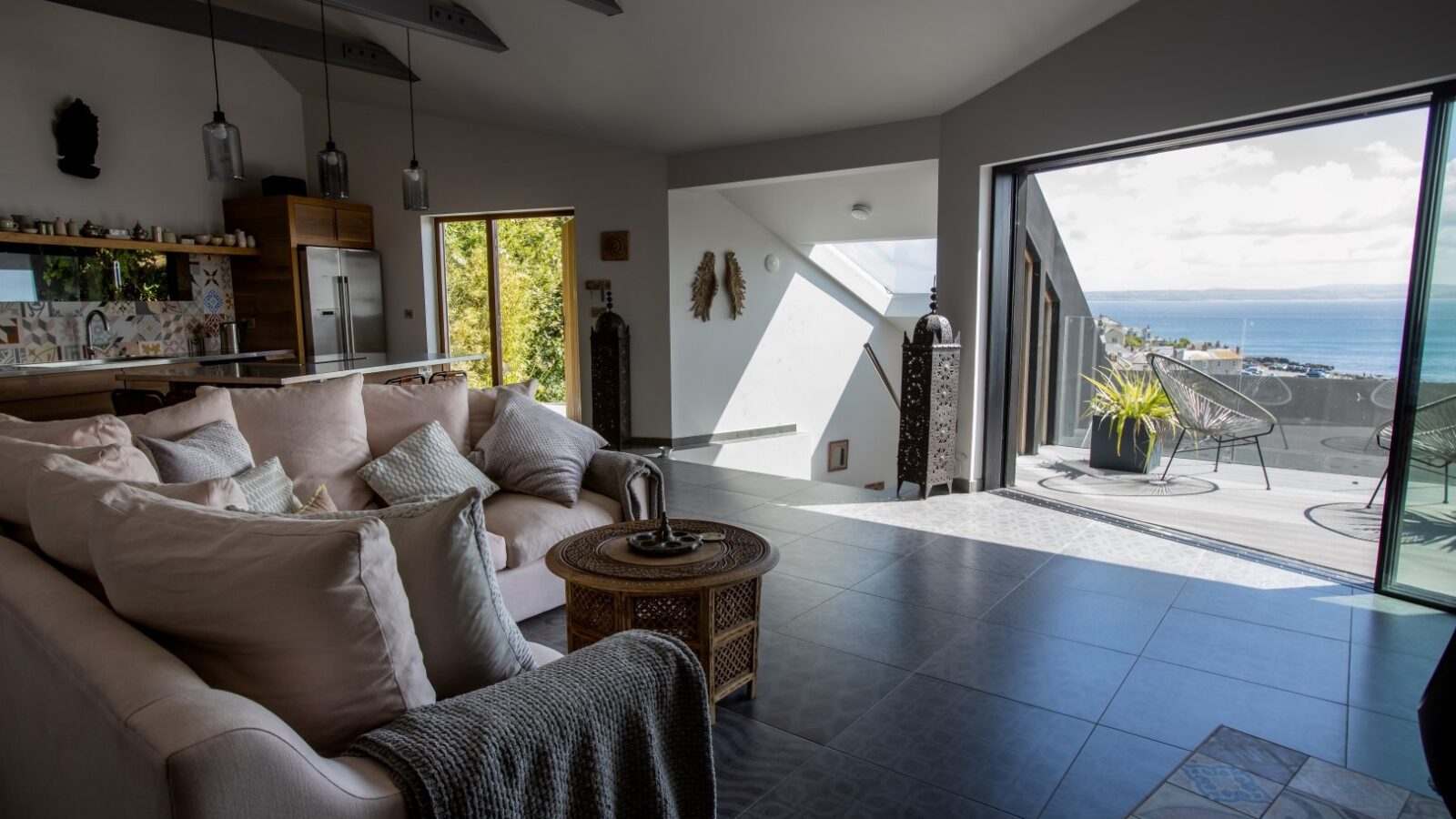 A modern, open-plan living room with a cozy beige sofa and a round wooden coffee table. The room features large floor tiles and opens to a spacious balcony with ocean views. Natural light floods the space through wide glass doors, creating a bright atmosphere that feels almost ethereal, like a fallen angel's sanctuary.