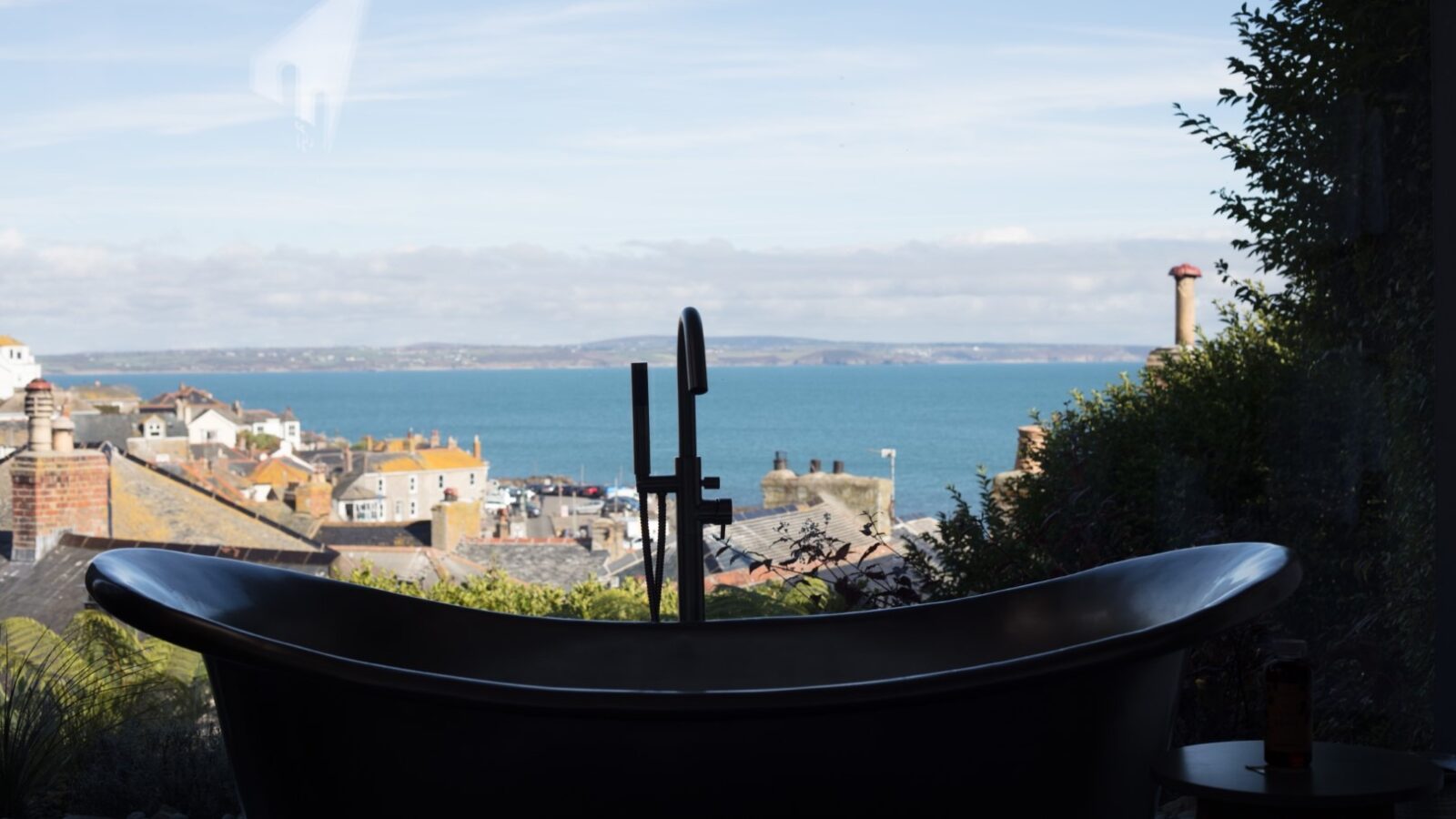 A sleek, modern bathtub is positioned in front of a large window with a stunning view of a coastal town and expansive ocean. The town's rooftops and chimneys lead the eye towards the calm blue sea and clear sky, creating a serene and picturesque scene that feels like it's been touched by a Fallen Angel.