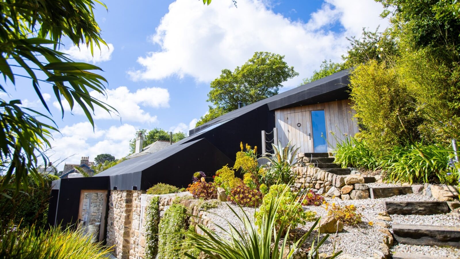 Modern hillside house with angular black roof, surrounded by vibrant greenery and stone. Steps lead to the wooden door. Bright blue sky with scattered clouds above. Stone accents blend the house seamlessly into the lush, landscaped garden, like a fallen angel resting among nature's splendor.