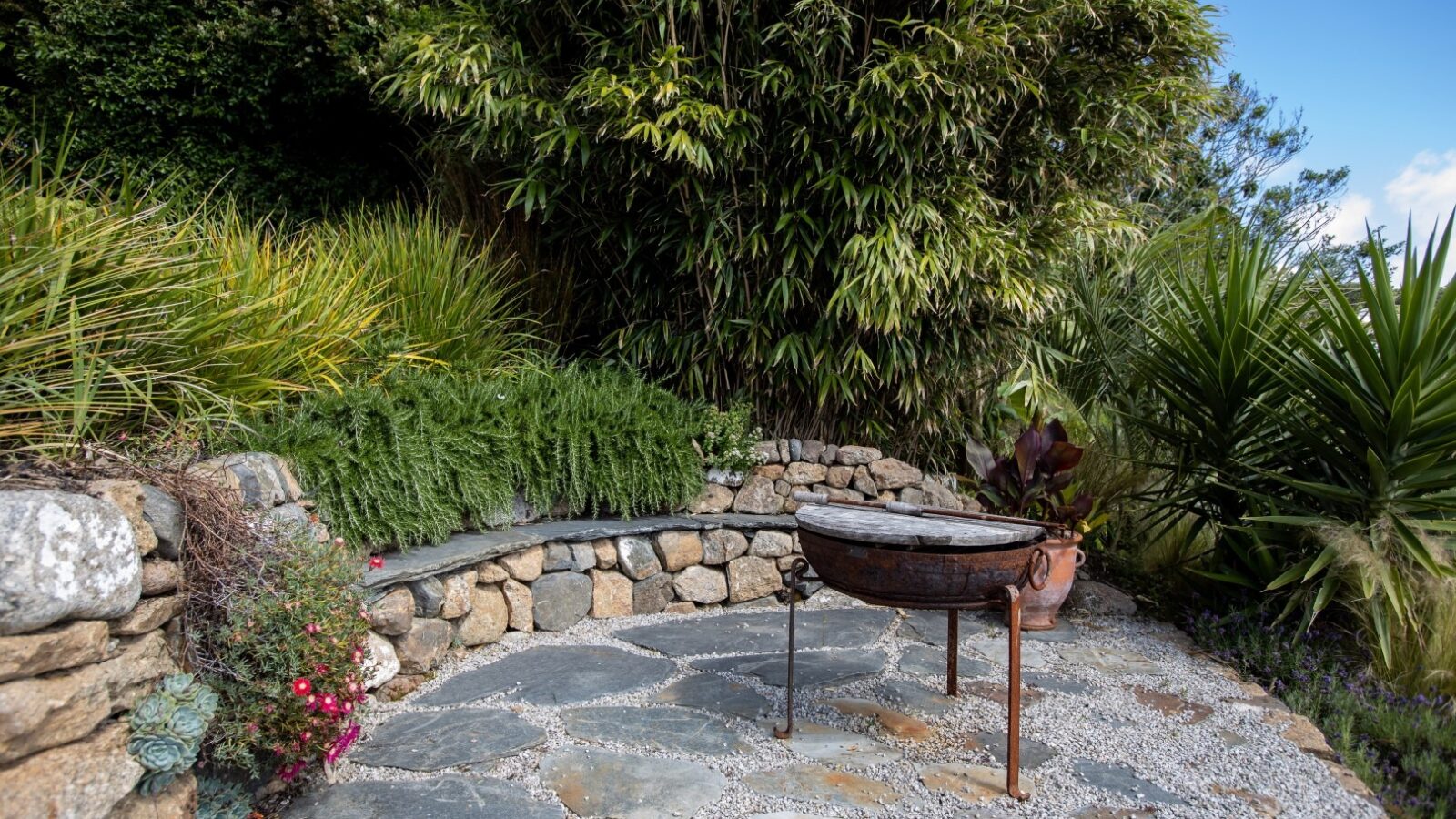 A tranquil garden corner features a curved stone bench set against a lush backdrop of green foliage and tall bamboo, creating a sanctuary that feels like the resting place of a fallen angel. In front of the bench stands a rustic metal fire pit on stone and gravel ground, with various plants and flowers adding to the serene ambiance.