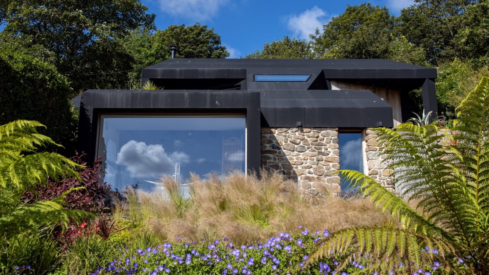 A modern two-story house with a black roof and stone walls, large windows on the lower floor, nestled among lush greenery like a fallen angel. The garden features ferns, tall grass, and vibrant purple flowers under a blue sky with scattered clouds.