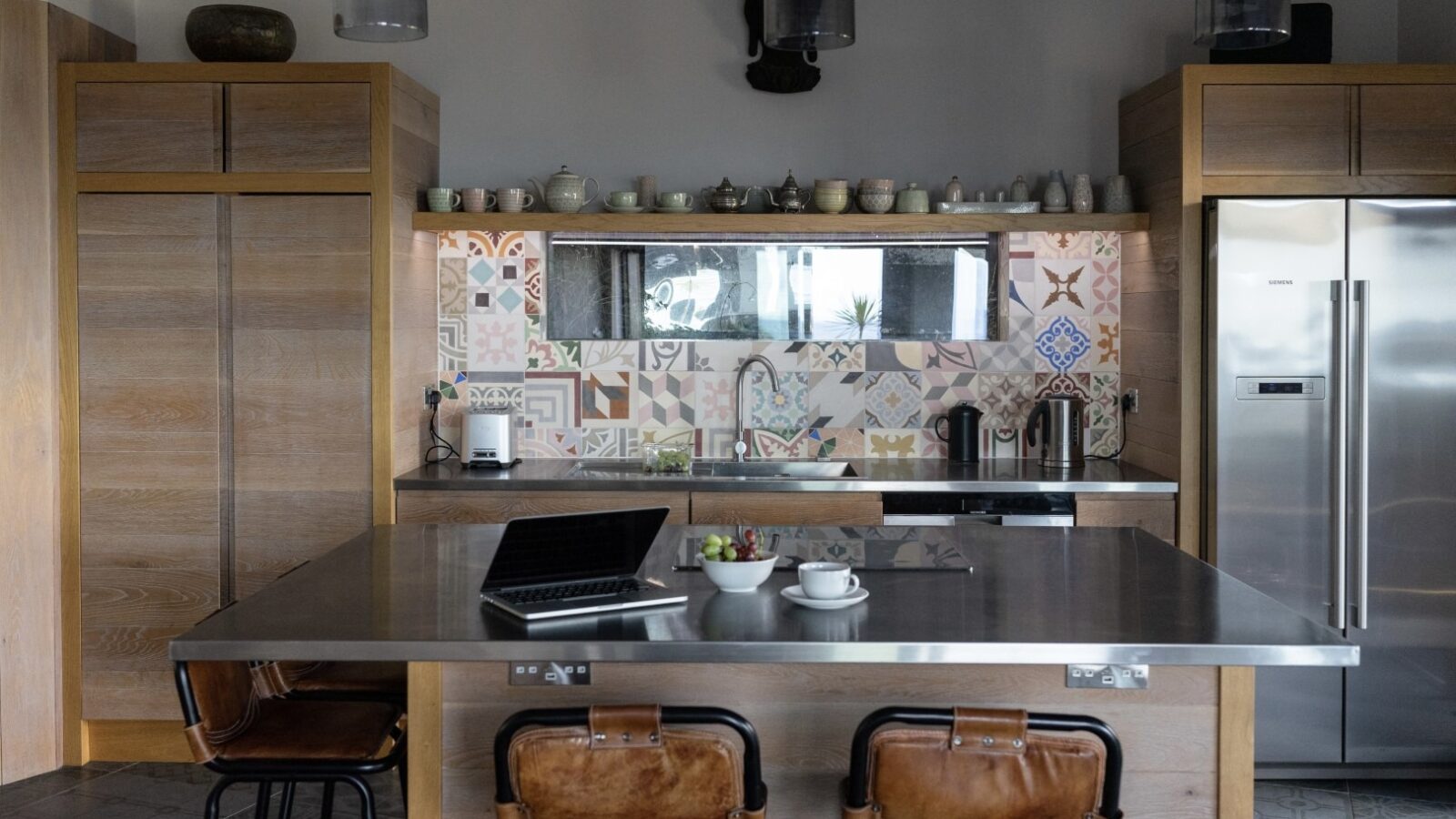 Modern kitchen with wooden cabinets, a stainless steel refrigerator, and a central island with a metal countertop. The island, reminiscent of a fallen angel in its elegance, has two leather stools and holds a laptop, a cup of coffee, and a fruit plate. The backsplash features colorful geometric tiles.