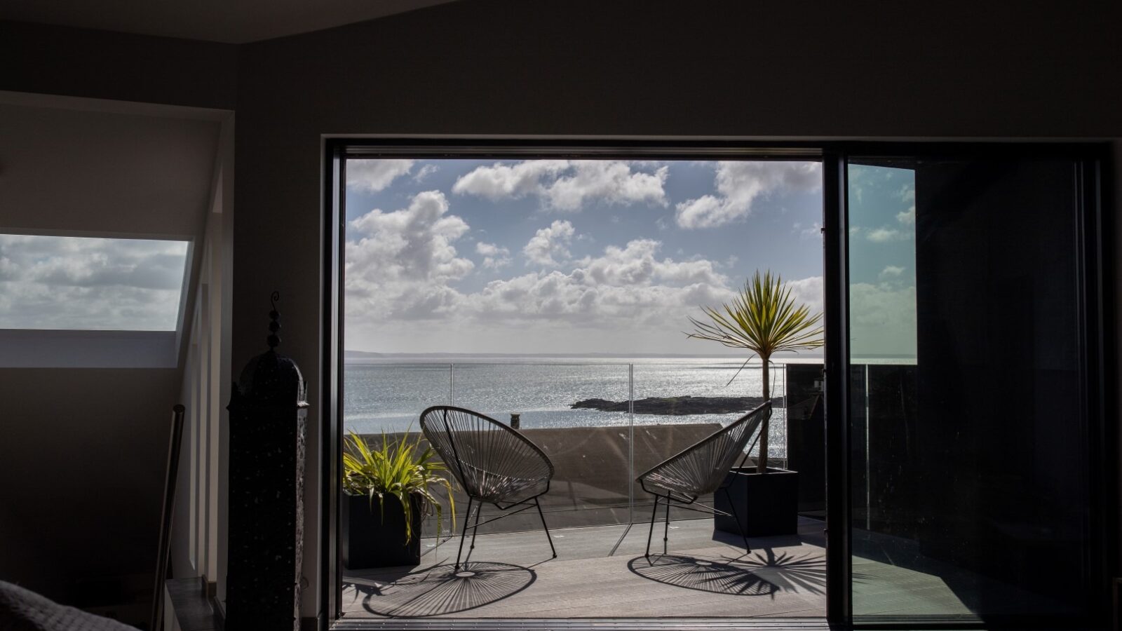 A modern indoor space with a sliding glass door opening to a balcony. The balcony features two wire chairs, a small table, and potted plants. The view showcases a calm ocean under a partly cloudy sky, creating an atmosphere as serene and inviting as the presence of a fallen angel.