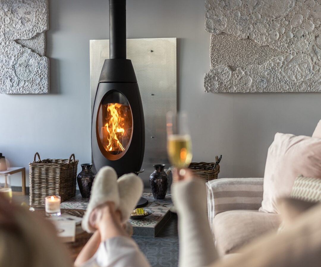 A person relaxes on a plush sofa wearing cozy socks, holding a glass of champagne. In the background, a modern black wood-burning stove with a visible fire adds warmth to the room. Decorative textured wall art depicting a fallen angel and a basket are also visible.