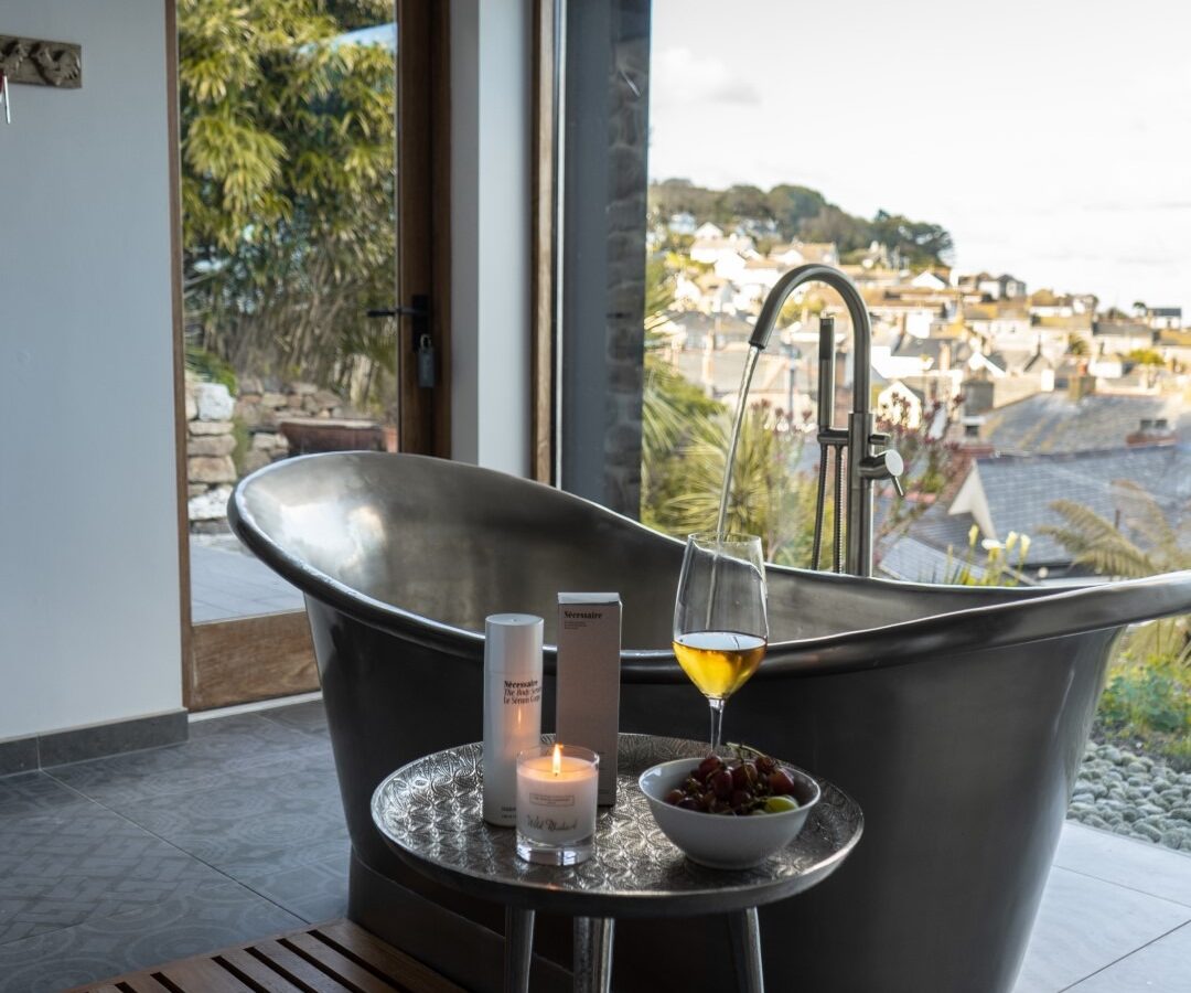 A modern bathroom features a sleek, freestanding metallic bathtub with a large window offering a scenic view of the town and greenery. Near the tub, reminiscent of a fallen angel's retreat, is a small table holding a lit candle, toiletries, a bowl of grapes, and a glass of yellow beverage.